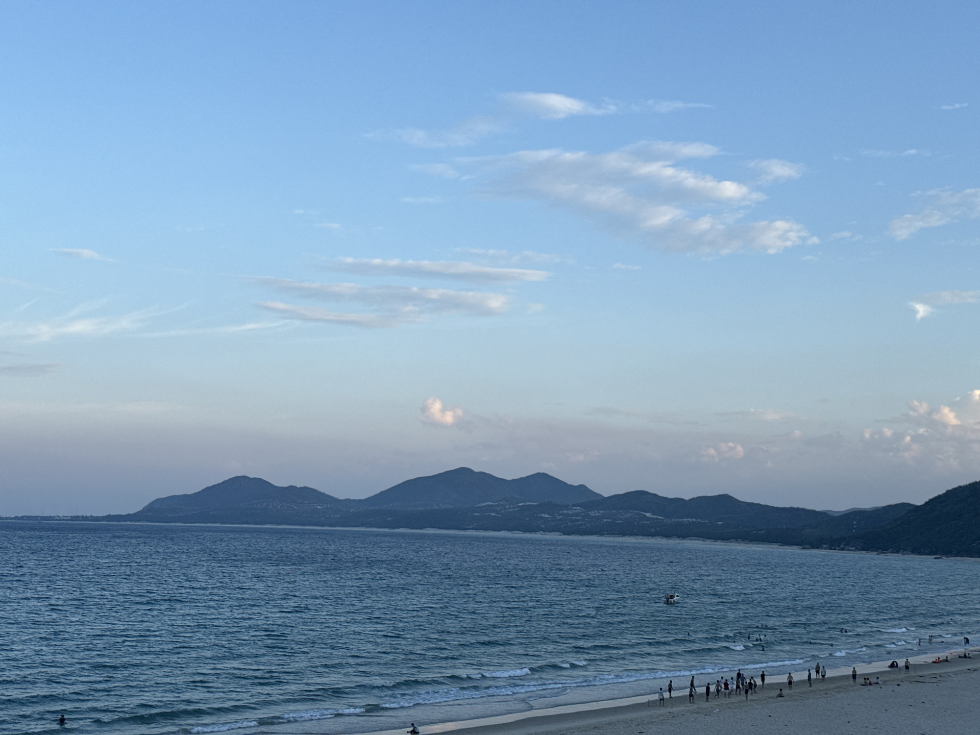 A panoramic view of a coastline with a beach, ocean, and mountains under a sky with scattered clouds. In the foreground, a sandy beach stretches across the bottom of the image, with a few people scattered along its expanse. Small waves gently lap at the shore. The ocean is a deep blue, with whitecaps visible in the distance. A small white boat is visible in the water. In the background, a range of dark, hazy mountains form a backdrop to the scene, with a lighter, hazy horizon line connecting them to the sea. The sky is a pale blue, with wispy white clouds scattered across it, some appearing elongated and streaky. The lighting suggests it is either early morning or late afternoon.