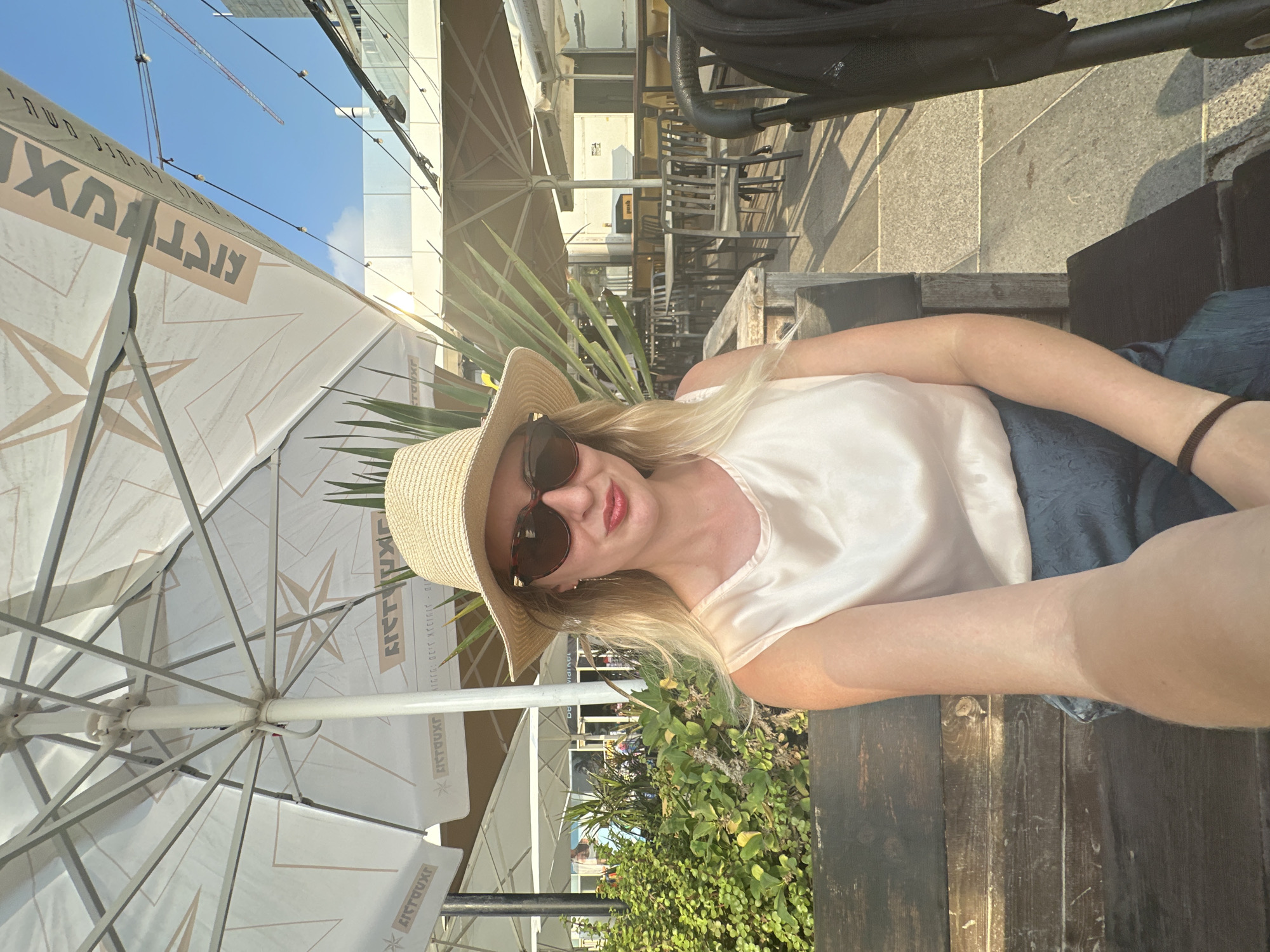 A woman with blonde hair and sunglasses, wearing a straw hat, a light-colored top, and a blue skirt, sits on a wooden bench. She is holding a phone in her left hand, taking a selfie. In the background, there are outdoor tables and chairs, umbrellas, and greenery. The scene appears to be an outdoor cafe in Bat Yam, Israel, with a view of the sky and some buildings in the distance. The overall lighting suggests it is daytime, possibly late afternoon, given the warm light and shadows.