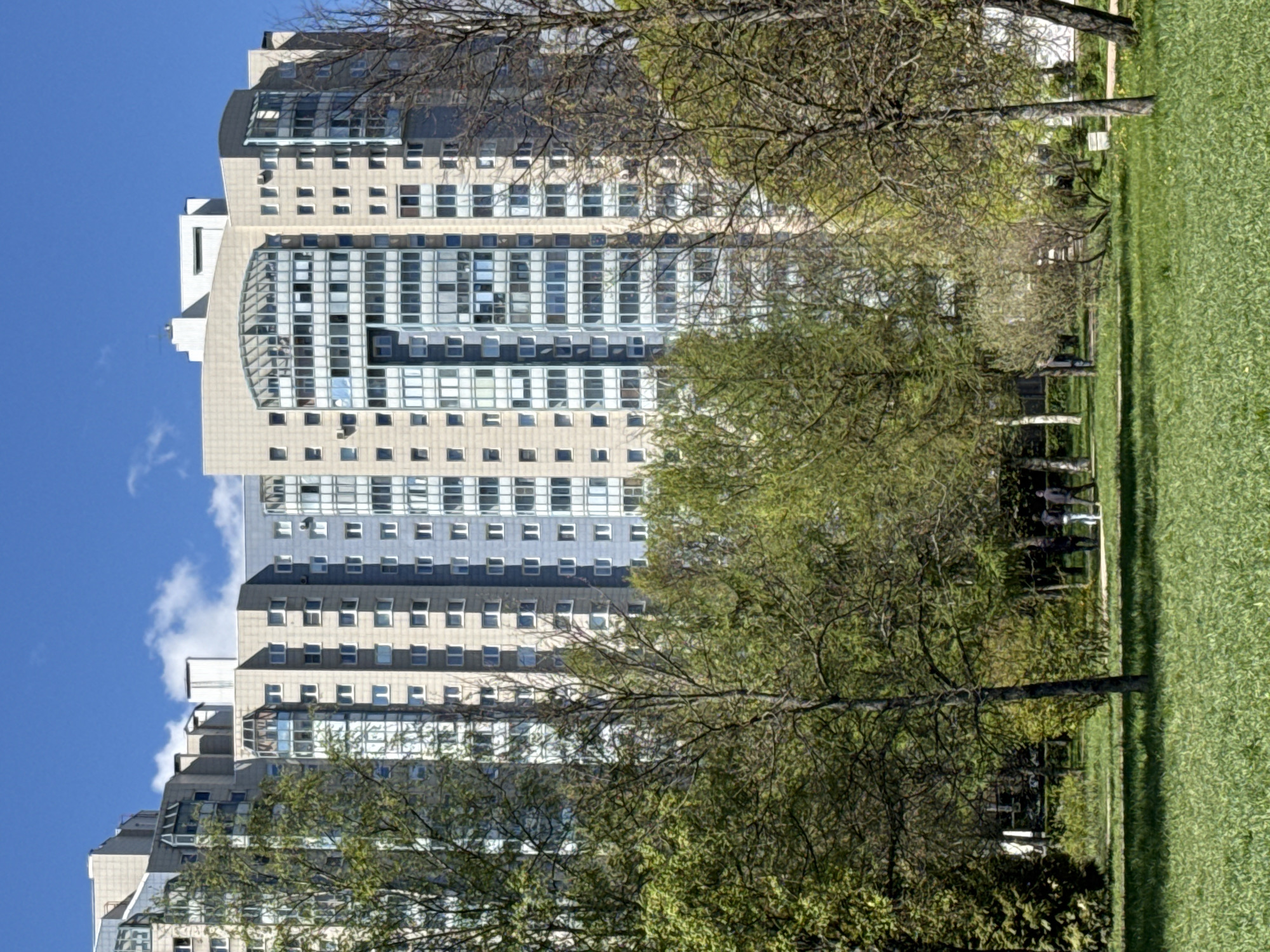 A vertical, eye-level shot depicts a tall, beige and grey building rising above a lush green park in Vsevolozhsk, Russia, under a clear blue sky. The building is rectangular with numerous small, square windows, and several levels feature curved, glass-enclosed areas. In the foreground, trees with budding green leaves partially obscure the building. A strip of green grass runs along the right side of the image, and some people are visible in the background, standing along the tree line.