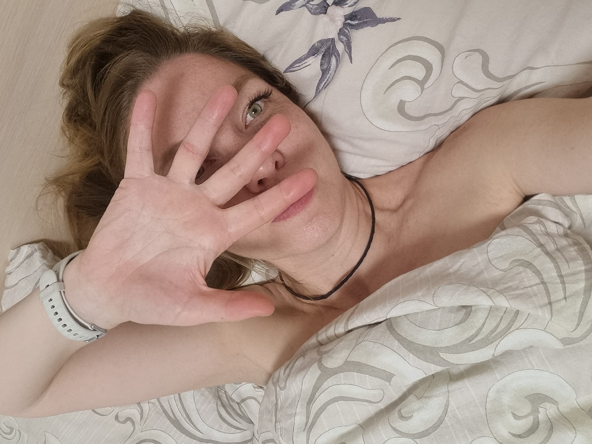 A woman is lying in bed, partially covered by a duvet with a grey and white pattern. Her head is on a pillow with a floral design. She has light brown hair and is wearing a thin black necklace. She is holding her hand up in front of her face, obscuring it. A white watch is visible on her left wrist. The background appears to be a pale beige or light wood-colored wall. The location is Chaikovsky, Russia.