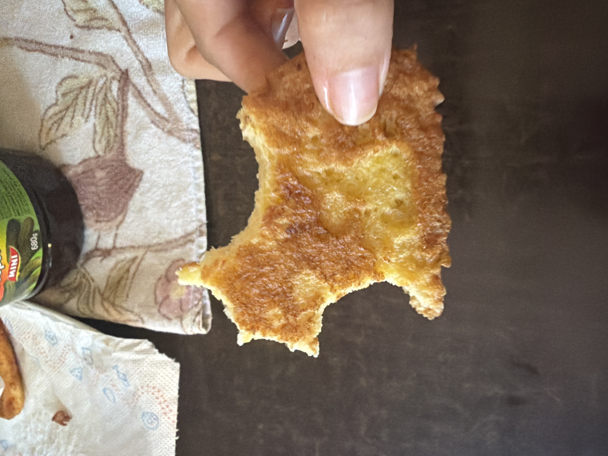 A person's hand is holding a piece of golden-brown fried bread against a dark brown surface. The bread has a porous texture and appears to be slightly curved, with a bite taken out of its left side. In the background, a portion of a white paper napkin with blue circular patterns and a piece of a patterned fabric with floral designs are visible. There is also a dark jar with a green label and white text that reads "MINI" and "680g". The overall scene suggests a casual setting, possibly a meal or snack. The specific location in Dromolaxia - Meneou Municipality, Cyprus cannot be determined from the visual information provided.