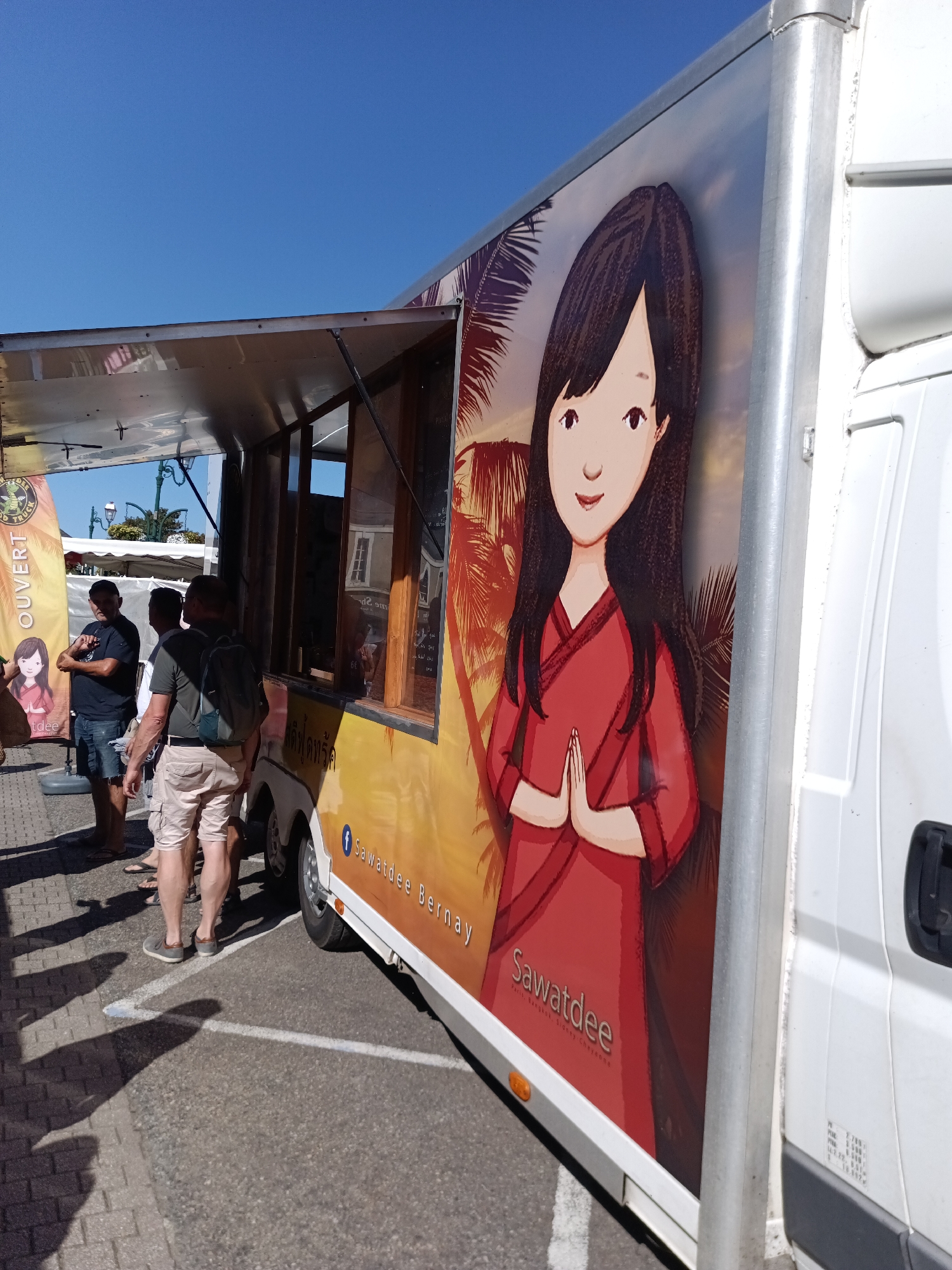 A food truck with a mural of a cartoon woman in a red dress is parked on a street in Le Neubourg, France, under a clear blue sky. The food truck is white and silver, with a serving window open. The mural depicts a woman with dark hair and a welcoming expression, her hands pressed together in a traditional greeting. Text on the truck reads "Sawatdee" and "Sawatdee Bernay", suggesting it is a Thai restaurant. Several men are standing near the truck, possibly customers. Shadows of the men are cast on the paved ground. In the background, there are buildings and streetlights.
