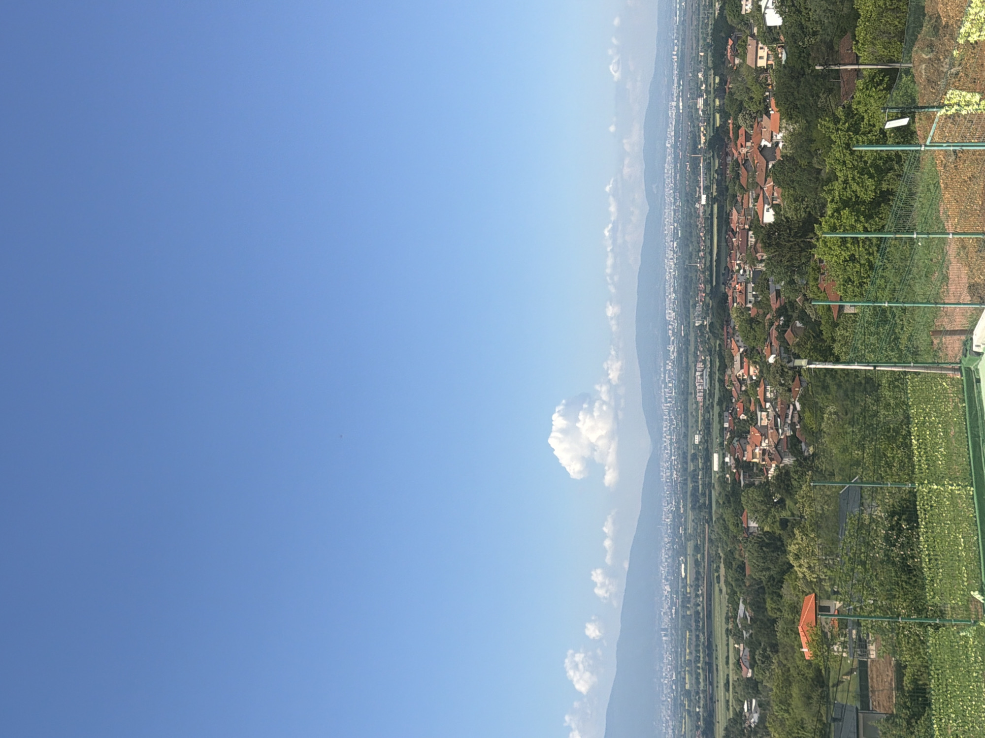 The image depicts a landscape in Voynegovtsi, Bulgaria, under a bright blue sky with scattered cumulus clouds. Mountains form a backdrop to the town. The town itself consists of buildings with predominantly red-tiled roofs interspersed with lush greenery. A green mesh fence and vegetation in the foreground suggest a residential or agricultural area.