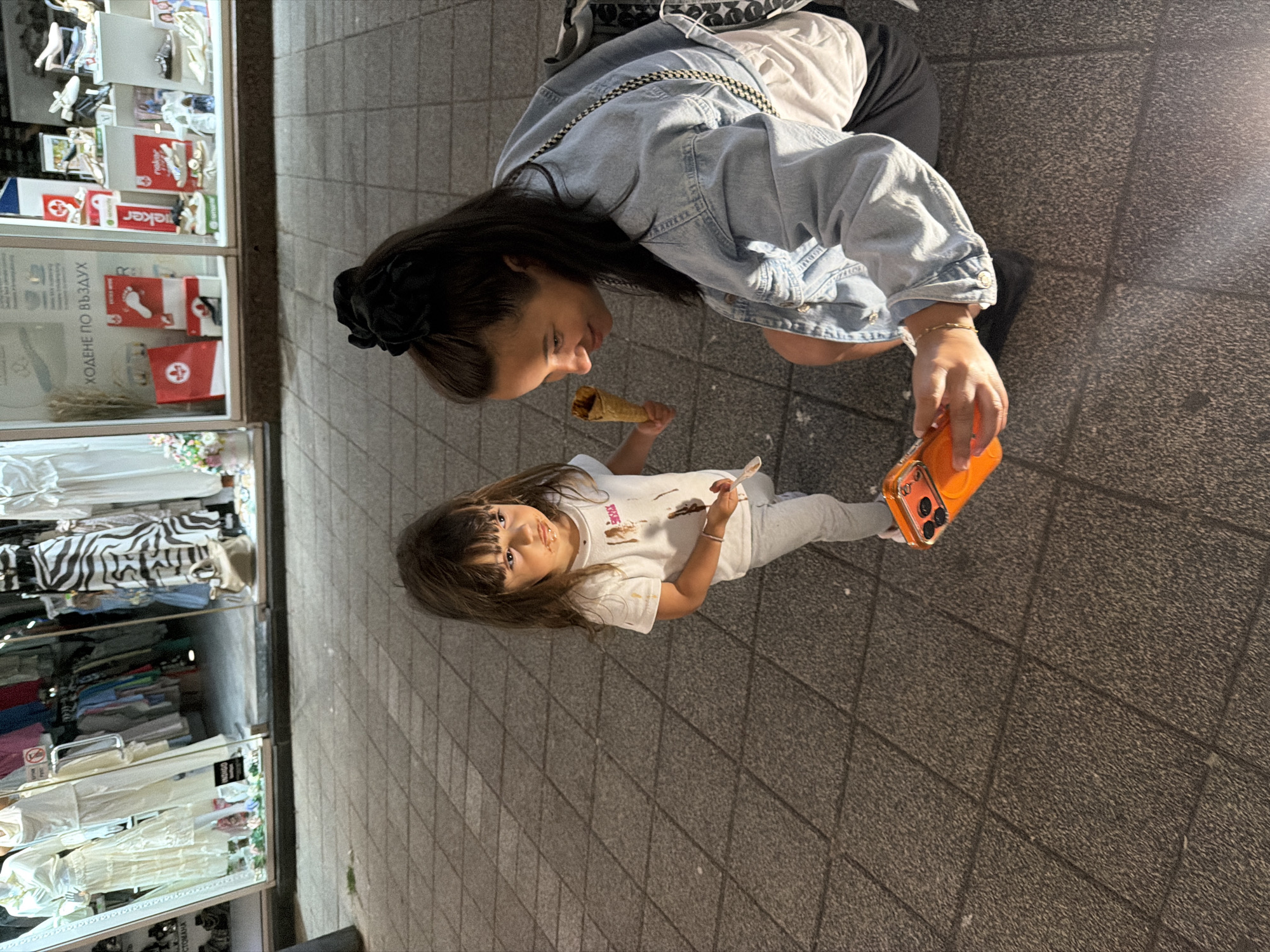 A young girl with long, dark, wavy hair is standing on a paved sidewalk, looking to her right with chocolate smeared around her mouth. She is wearing a white t-shirt with brown smudges and light grey pants. She holds a spoon in her right hand. To her right, a woman with dark hair pulled back in a black scrunchie is squatting down, looking at the girl and holding a smartphone in her right hand. The woman is wearing a light blue denim jacket over a white t-shirt and black shorts. She has a black crossbody bag with a patterned strap. The background shows storefronts displaying clothing and shoes. The setting is likely a commercial street in Burgas, Bulgaria, given the context. The time of day appears to be evening, as indicated by the lighting.