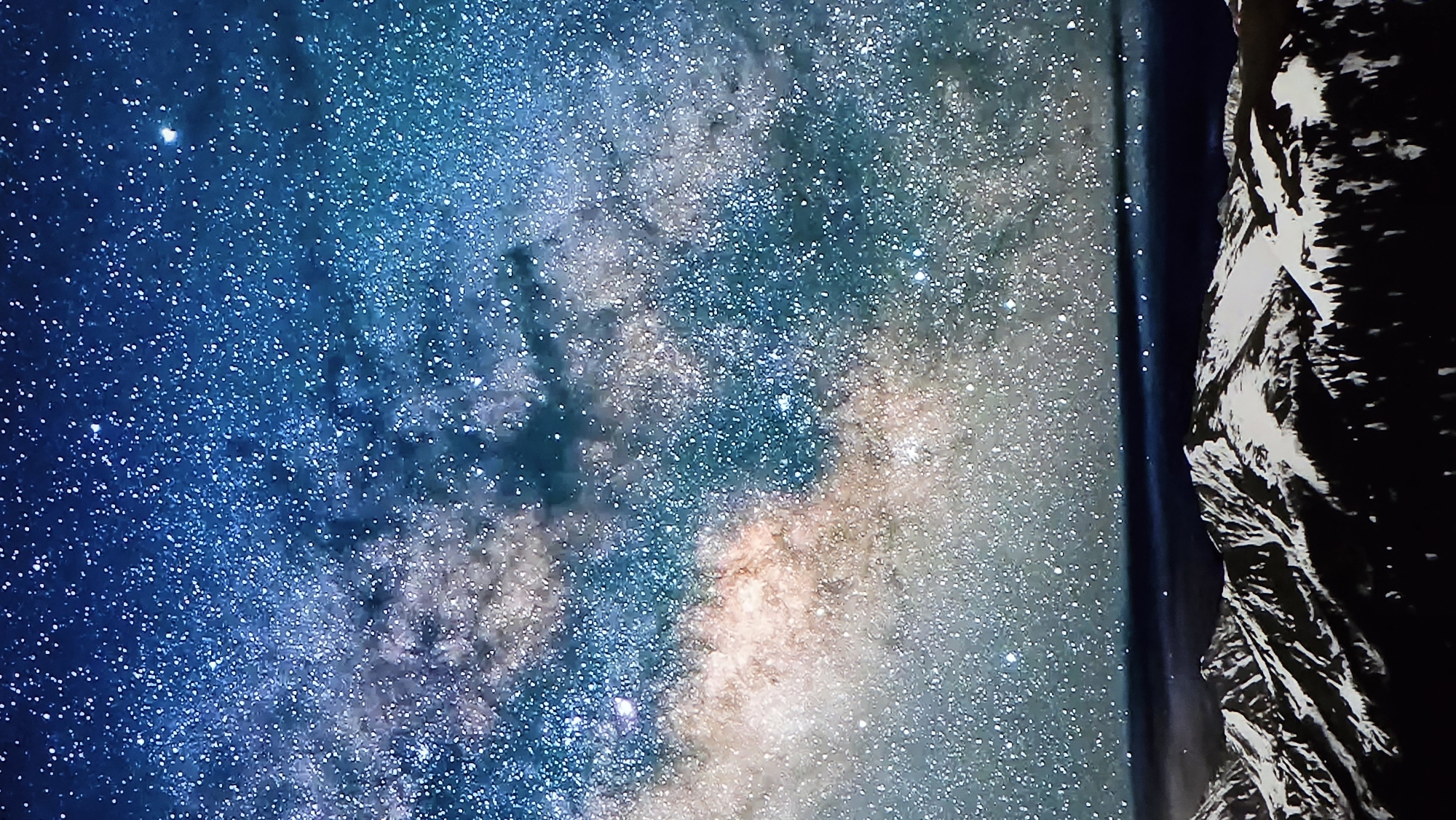 The image depicts a breathtaking view of the Milky Way galaxy against a dark night sky, with a rugged, mountainous terrain dominating the right side of the frame. The galaxy is a swirling expanse of stars and nebulae, with vibrant blues, purples, and hints of orange and pink, creating a sense of immense depth and cosmic wonder. The stars are densely packed, some appearing as bright pinpricks of light, while others are more diffuse, forming cloudy bands.
The mountainous foreground is rendered in stark black and white, with sharp, angular forms suggesting jagged peaks and cliffs. The contrast between the celestial display and the earthly landscape is dramatic, evoking a feeling of awe and insignificance in the face of the universe's vastness.
The time of day is clearly nighttime, as evidenced by the presence of stars and the dark sky. There are no discernible weather conditions, but the clarity of the stars suggests a clear atmosphere. The location cues are not evident within the image itself, but the provided context of Dobrich, Bulgaria, places this scene within that geographical region.
No people or specific objects are visible, making the focus entirely on the natural phenomena of the night sky and the terrestrial landscape. The overall mood conveyed is one of profound beauty, mystery, and the sublime. There is no visible text in the image.