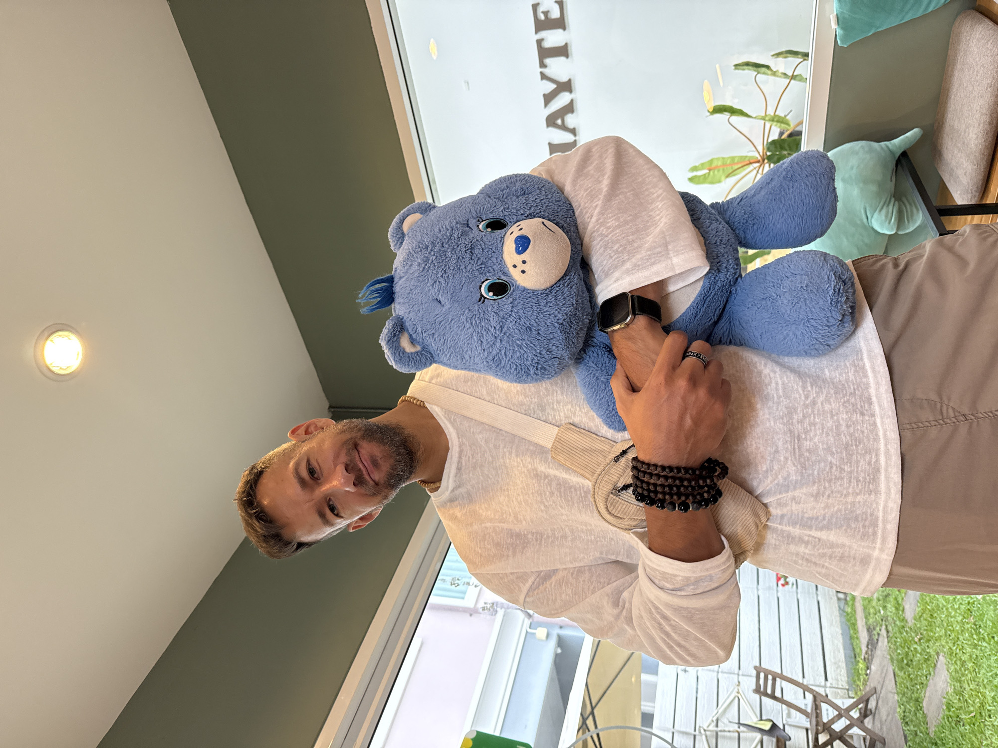An adult male of Caucasian appearance, with short, dark hair and a goatee, is pictured indoors, holding a large, blue plush bear. He is wearing a white, long-sleeved shirt, tan shorts, and several beaded bracelets on his left wrist. A light-colored crossbody bag is worn over his right shoulder. The male is smiling and looking at the camera. The plush bear has blue fur, big eyes, a blue heart-shaped nose, and blue tufts of hair on its head. In the background, a window reveals an outdoor scene with greenery and outdoor seating, suggesting a cafe or restaurant setting. The wall behind the man is painted green. A sign with the word "HAYTE" is visible through the window, but is partially obscured. The overall ambiance suggests a casual and friendly environment. The scene is consistent with a cafe in Bangkok, Thailand, given the tropical vegetation visible outside and the modern interior design.