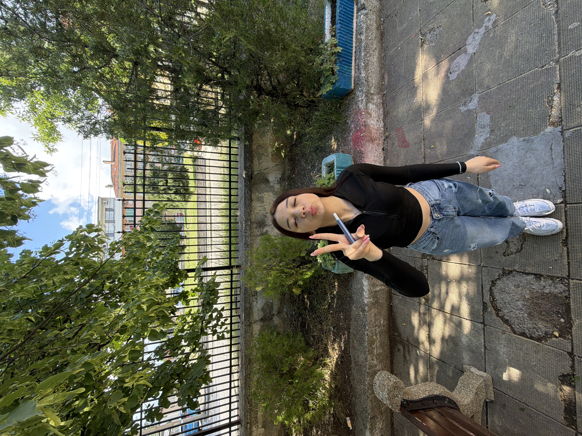 A young woman is captured posing outdoors on a bright, sunny day in what appears to be an urban or suburban setting in Bobov Dol, Bulgaria. She stands on a paved sidewalk, looking towards the camera with a playful expression, making a pouty face and giving a peace sign with her right hand while holding a slender, pen-like electronic device. She has long, dark hair and is dressed in a black, long-sleeved crop top, ripped light-wash blue jeans, and white sneakers.
The background features a black metal fence running horizontally behind her, separating the paved area from a patch of greenery with small trees and bushes. Beyond the fence, several multi-story buildings with red-tiled roofs are visible under a clear blue sky dotted with white clouds. Large, lush green trees with dense foliage frame the upper and left parts of the image, casting dappled sunlight and shadows on the ground. A stone bench is partially visible in the foreground to the right. The paved ground shows some wear and patches of repair. The overall scene conveys a casual, relaxed atmosphere in a daytime setting. No visible text is present in the image.