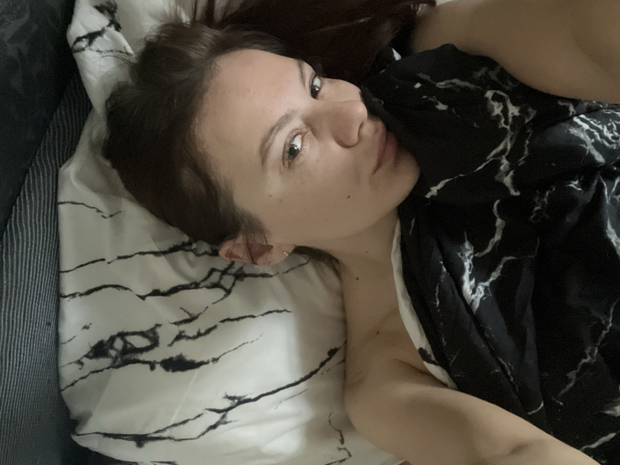 A woman is lying in bed. She has brown hair and fair skin. She is looking at the camera with a neutral expression. She is lying on a pillow with a white and black marble-like pattern. She is covered with a black and white marble-like blanket. There is a dark grey object in the upper left corner, possibly a couch. The image was likely taken indoors. The location is not possible to determine based on visual information.