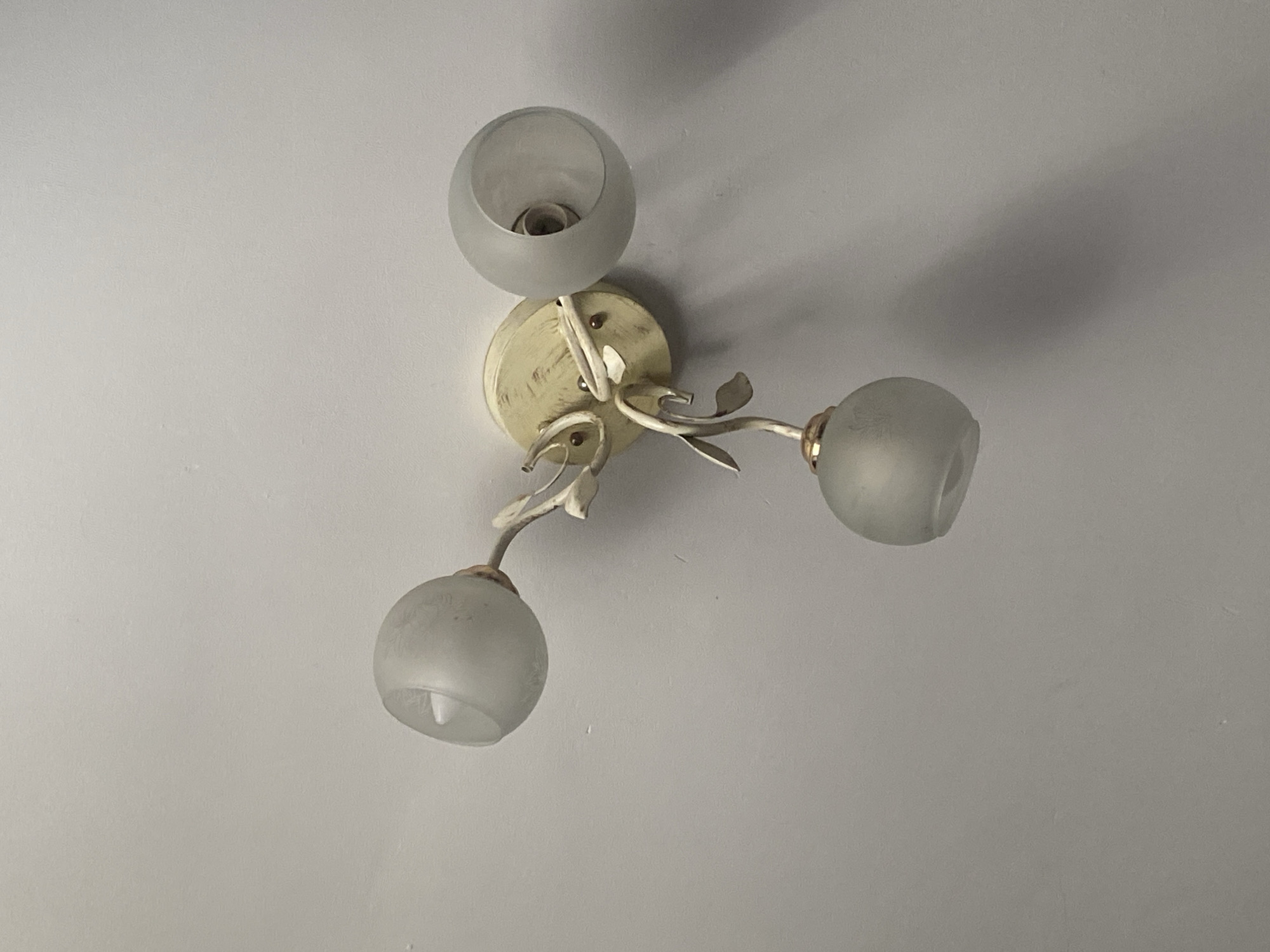 The image shows a close-up, overhead view of a ceiling light fixture. The fixture is cream-colored with three frosted glass globes, each housing a light bulb. The metal arms of the fixture are shaped like branches with small leaf accents. The ceiling is a plain, textured white. The scene is indoors, and there is no indication of the specific location in Keŋeş, Kyrgyzstan, other than it being a part of a building. There are no people or actions depicted.