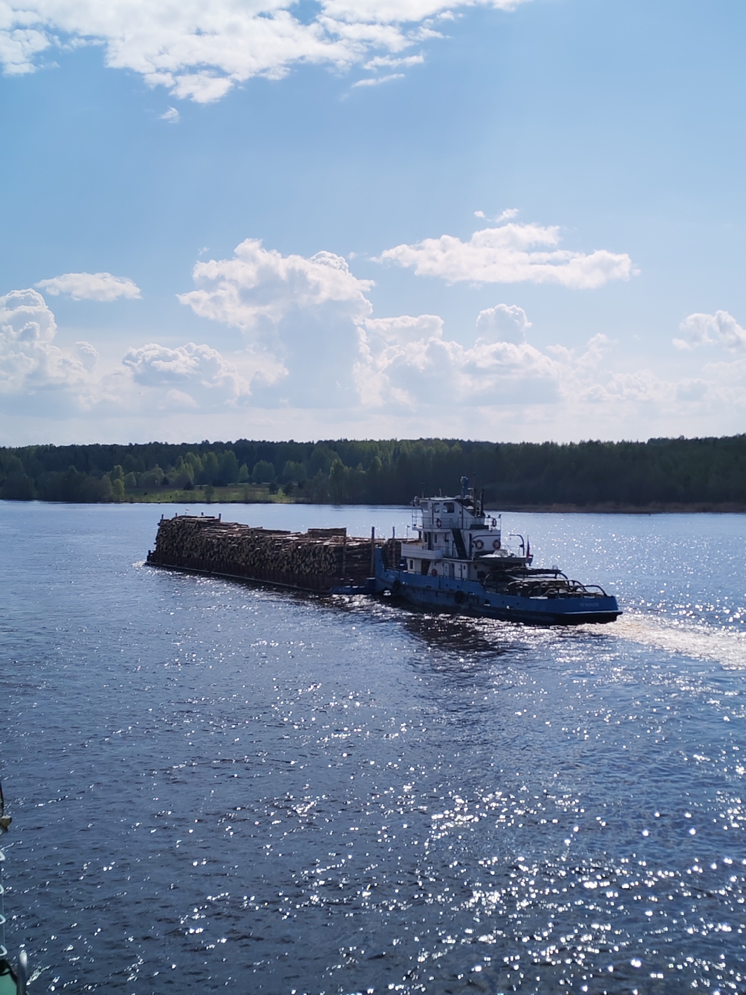 A barge laden with timber logs is being towed along a waterway by a tugboat in the city of Popovo, Russia. The water surface reflects sunlight. A green forest is visible in the background along the shoreline. The sky has blue patches and white clouds.