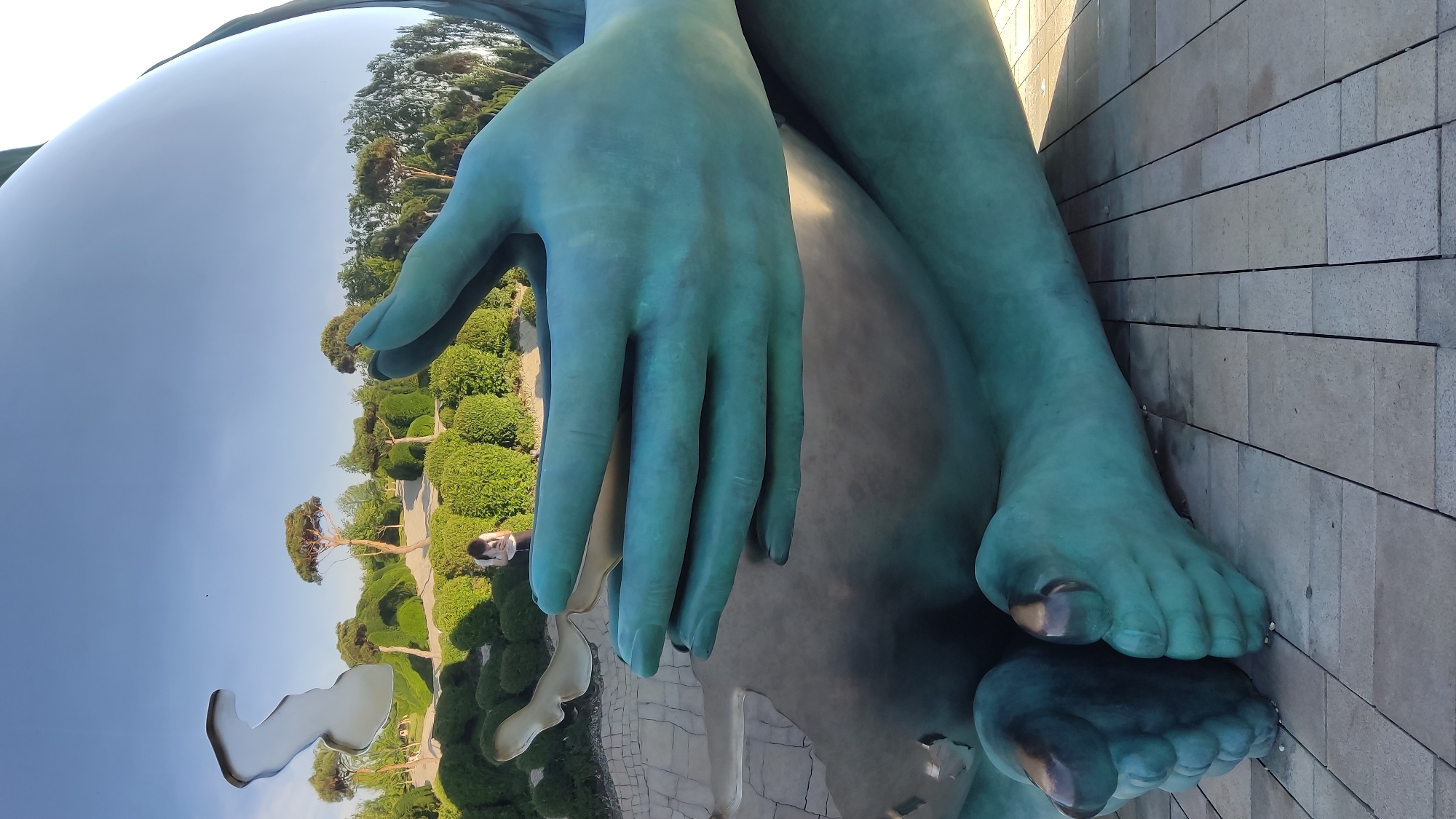The image shows a large, reflective sculpture of a person's hands and feet, made of a greenish-blue metal. The sculpture is located outdoors, on a paved area made of rectangular stones. In the background, there is a park-like setting with manicured bushes and trees. A person, with dark curly hair and wearing a white top and dark bottoms, is visible in the reflection of the sculpture, looking at a phone. This sculpture is the "World" monument located in Krasnodar, Russia.