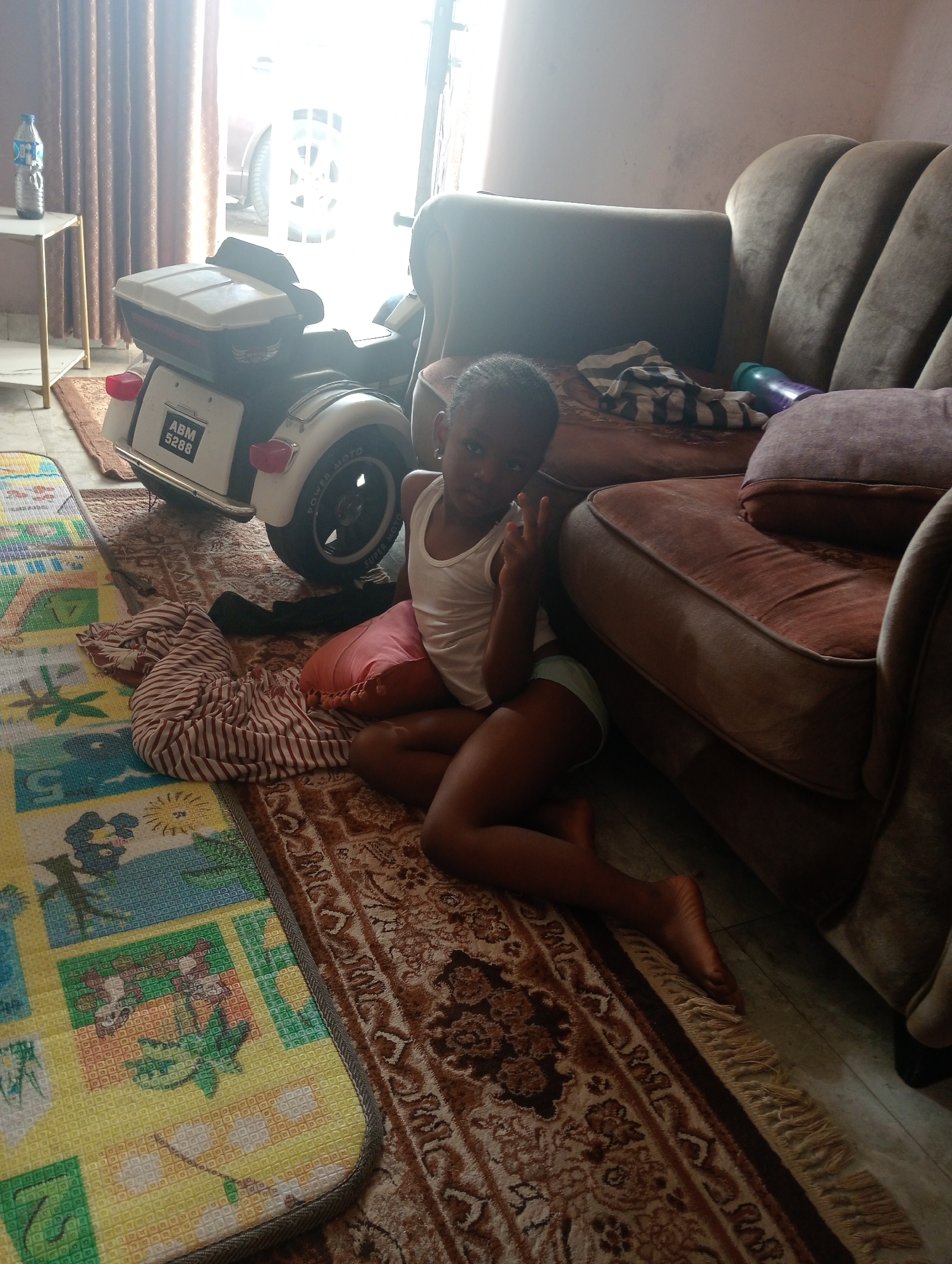 A young girl is sitting cross-legged on a patterned rug next to a brown sofa. She is wearing a white tank top and light-colored shorts, and is holding up two fingers in a peace sign. Behind her is a toy motorcycle, and to the left is a colorful playmat. A table with a water bottle sits in the background near a window. The location is Jos, Nigeria.