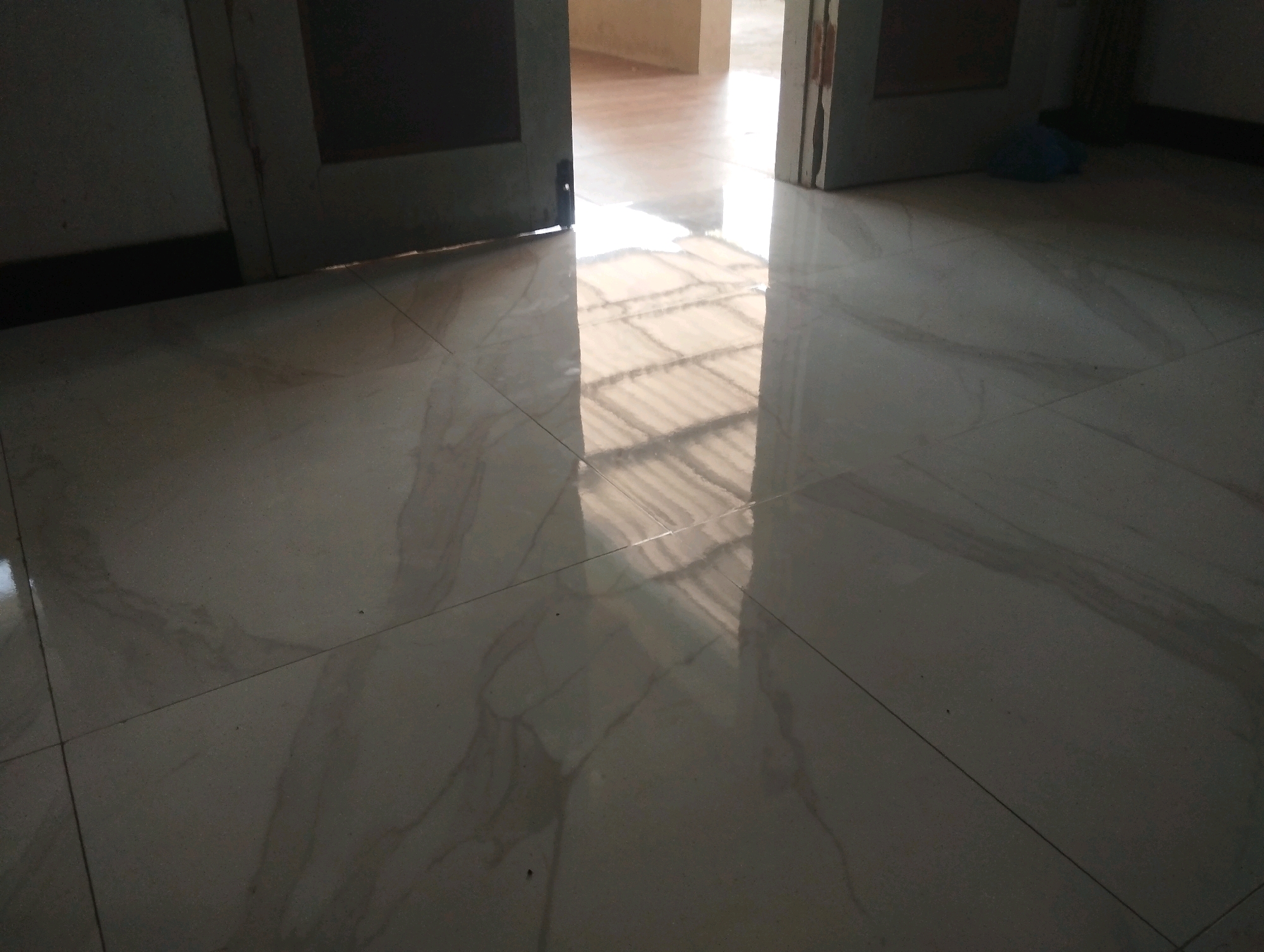 The image shows a tiled floor in what appears to be a room with open doorways. The floor is made of large, light-colored tiles with subtle veining, suggesting a modern or well-maintained interior. Light streams in from an adjacent area, creating a bright reflection on the floor, which hints at a ceiling or structure with a repeating pattern, possibly corrugated metal or blinds. Two doorways are visible, one on the left and one on the right. The doorway on the left is partially closed, revealing a dark interior, while the doorway on the right is fully open, showing a well-lit, spacious area beyond. In the background, near the right doorway, a crumpled blue object, possibly a plastic bag, rests on the floor. The overall impression is of an indoor space with natural light entering from an adjoining room, possibly an office, hallway, or a residential area. The scene is clean and uncluttered, with the focus on the interplay of light and shadow on the polished floor. The location in Jatirejo, Indonesia, implies a tropical or subtropical climate, where such lighting conditions are common.