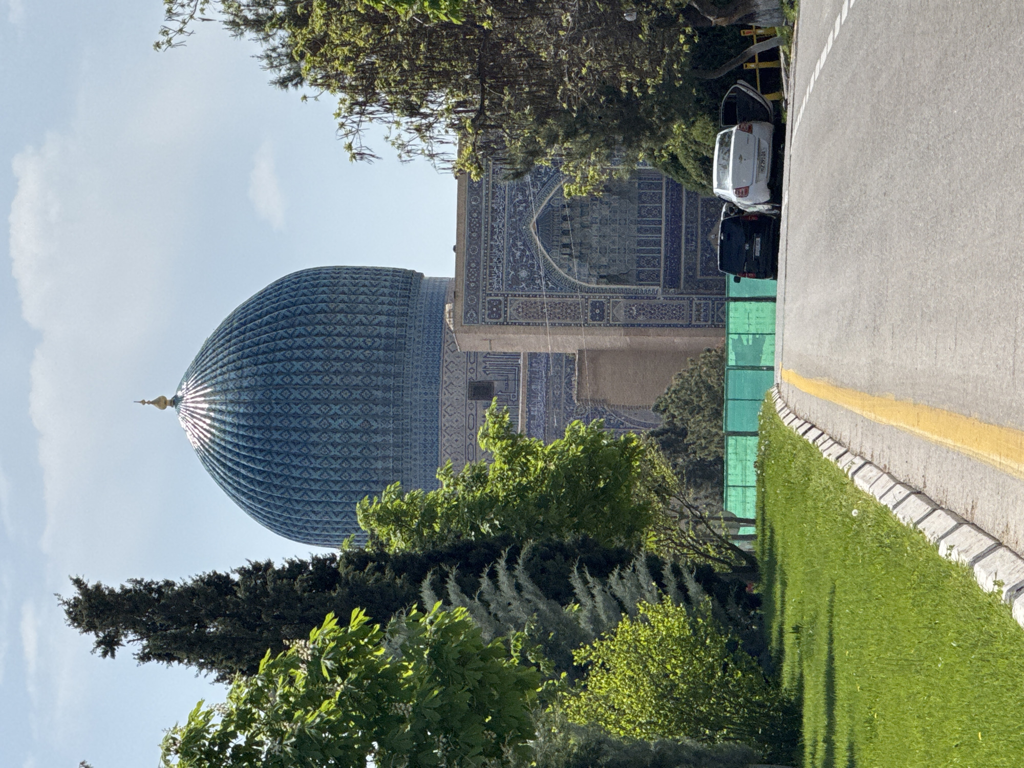 The image captures a view in Samarkand City, Uzbekistan, featuring the Gur-e-Amir Mausoleum. The mausoleum's large, ribbed, blue dome is partially visible behind trees. A section of the mausoleum's tiled facade is also shown. Next to a road with parked cars, there is grass, with tree and vegetation in the foreground, partially obscuring the architectural details. The sky is lightly clouded.