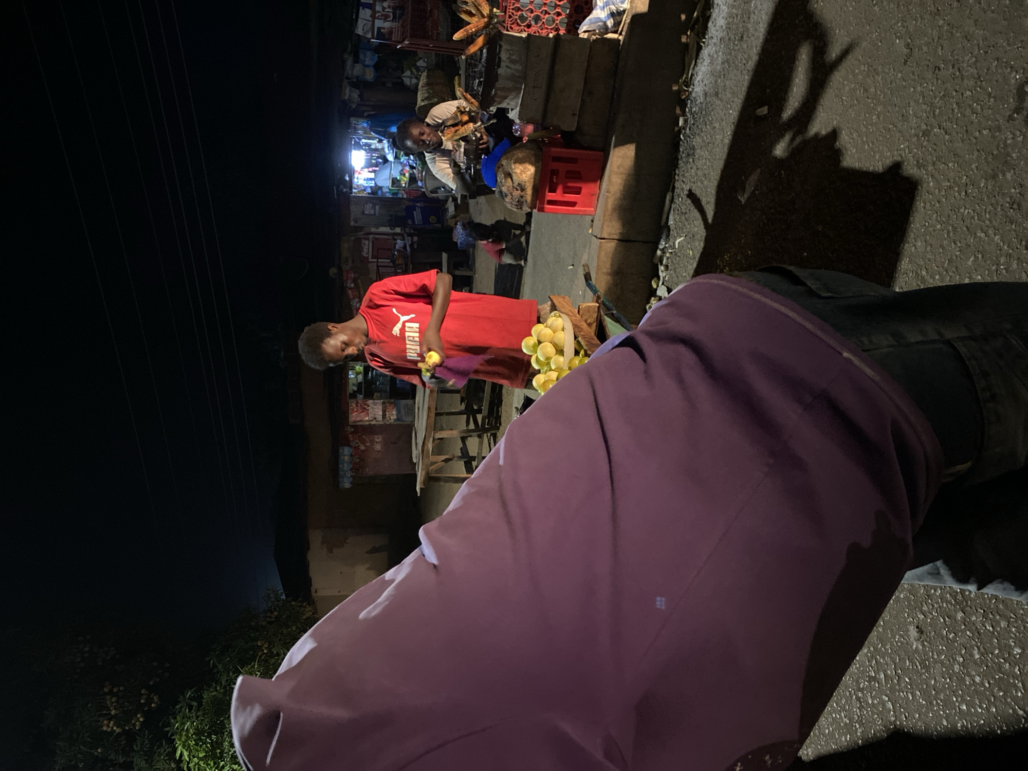 The scene depicts a bustling street in Bukuru, Nigeria, at night. A young man wearing a red Puma t-shirt is seen holding a yellow fruit (possibly a lemon or lime) at a fruit stand. The fruits are piled in a wooden container. In the background, another vendor is preparing corn on a makeshift table. Red plastic crates are used for storage. The street is lit by dim storefront lights. In the foreground, a person wearing a maroon jacket stands at the edge of the scene, partially obscuring the view. The road surface is made of asphalt and shows signs of wear. Shadows are prominent due to the limited lighting. Overhead power lines are visible against the dark sky.
