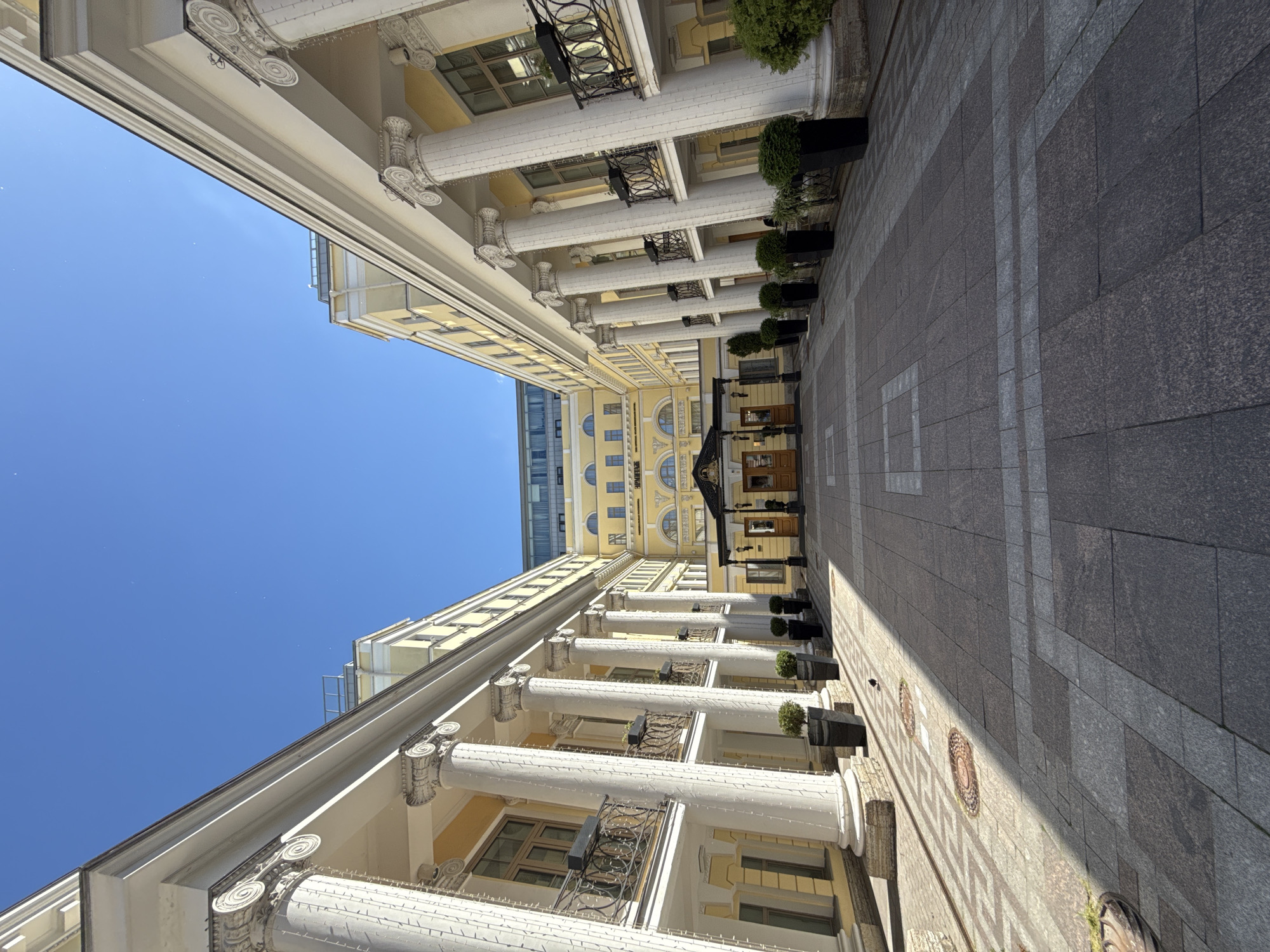 This image shows the courtyard of the Hermitage Museum in Saint Petersburg, Russia, on a clear, sunny day. The courtyard is paved with stone tiles and features classical architecture with tall white columns and yellow facades. Large potted plants are placed along the sides of the courtyard. In the background, there is a building with arched windows and a grand entrance. The sky is a bright, clear blue.