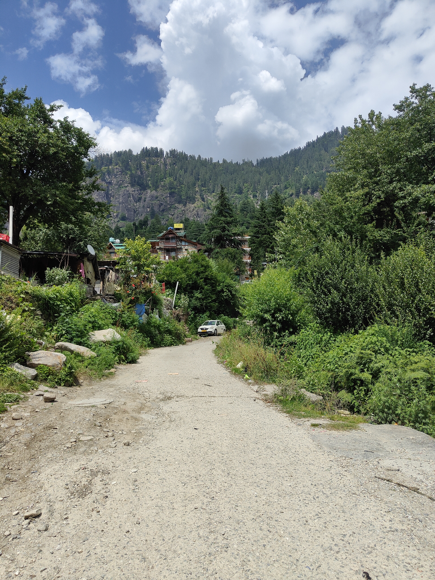 The image depicts a rural scene in Vashisht, India, with a narrow, unpaved road leading uphill. Lush green vegetation and trees line both sides of the road, with a steep, forested mountainside in the background. A few buildings, including what appears to be a guesthouse or hotel with a yellow sign, are visible amongst the trees. A white car is parked on the side of the road. The sky is partly cloudy with a mix of blue sky and white clouds. The overall impression is one of a remote, mountainous village.