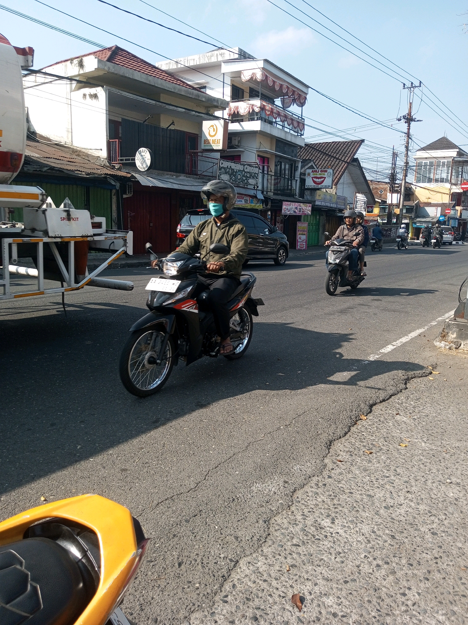 The image depicts a street scene in Wonosobo, Indonesia, likely in a commercial area, with a bright, sunny sky. In the foreground, a yellow motorcycle seat is visible, angled towards the left. Dominating the mid-ground, a man wearing a helmet and a light brown jacket is riding a black motorcycle. He is also wearing a blue face mask, and his vehicle has a license plate starting with "AA". To his right, another motorcycle with two occupants is also traveling. The background features a row of buildings with multiple stories, some displaying awnings and signs. One sign is for "NEST MEAT," and another depicts a smiling tooth, indicating a dental clinic. Power lines are strung across the sky, and a utility pole is visible. The road surface shows signs of wear and tear, with cracks and variations in texture. A white truck with "SPILL KIT" written on its side is parked on the left side of the road. The overall impression is of a typical day in a moderately busy urban environment.