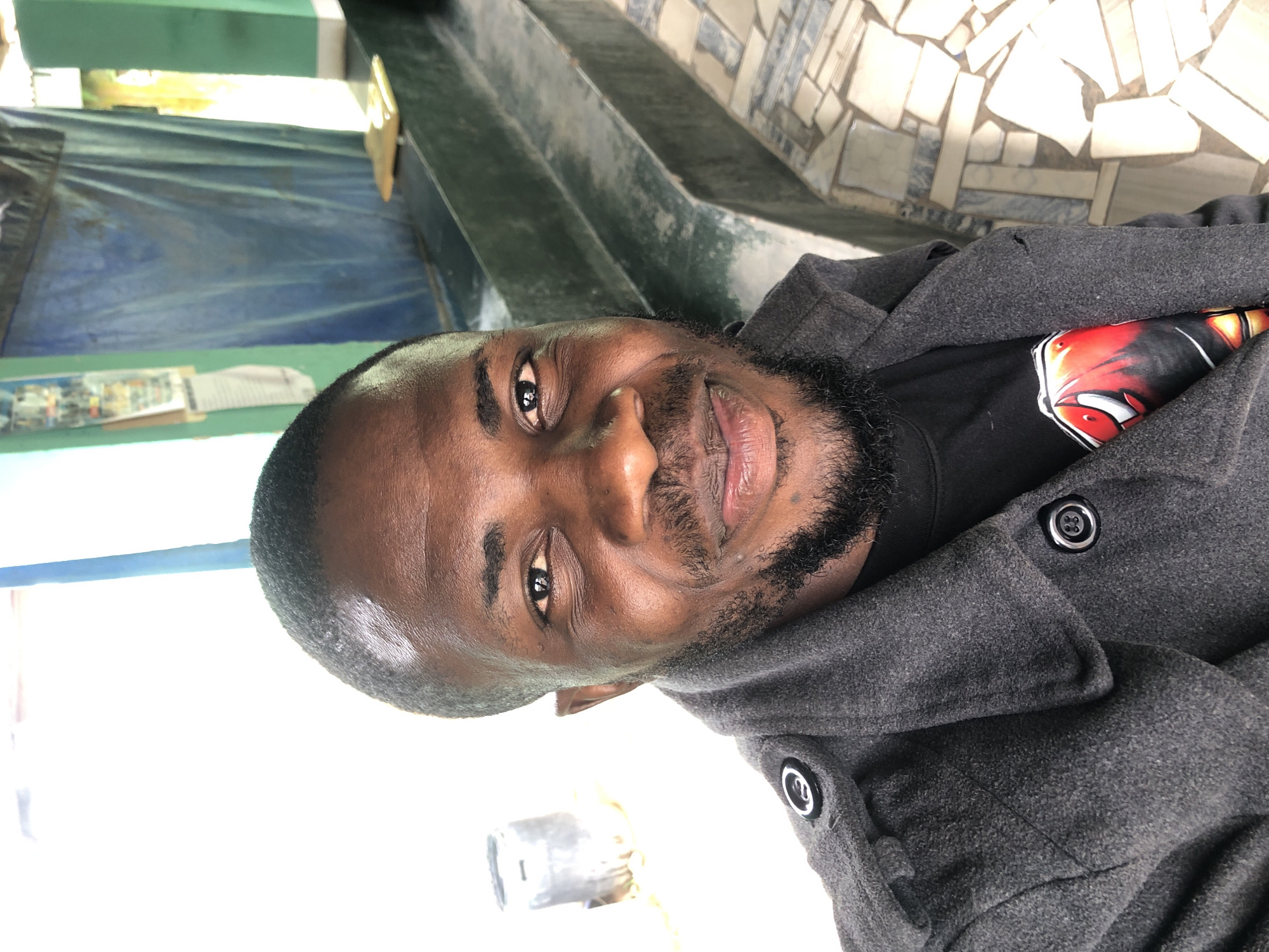 A young man with dark skin, a short beard, and dark, closely cropped hair is featured in a close-up portrait, looking directly at the camera with a slight, friendly smile. He is wearing a dark grey, possibly wool or felt, peacoat or heavy jacket with prominent black buttons. Underneath, a black t-shirt with a colorful graphic design, primarily featuring red, white, and black elements that are not clearly identifiable as text, is visible.
The setting appears to be outdoors or in a semi-open area, under bright daylight. In the background, to the man's left, there are green painted structures and a draped blue tarp or sheet. Further back and slightly to the right, a distinctive wall is visible, covered in light-colored, irregularly shaped mosaic tiles, suggesting a decorative or architectural feature. The lower right portion of the background is a bright, overexposed white area, possibly a bright sky or a highly illuminated wall. The overall activity is a simple pose for a photograph, capturing the man's relaxed and approachable expression. No legible text is visible in the image.