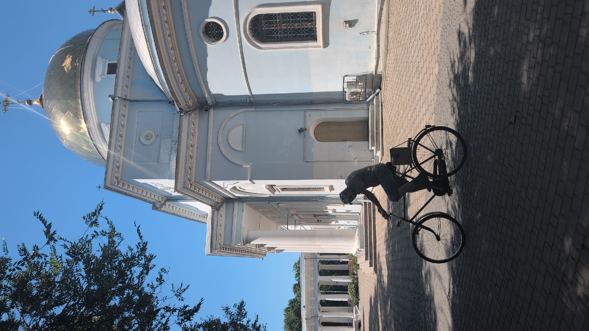 A person is riding a bicycle in the courtyard of a blue-colored church with a golden dome. The person is wearing a cap and a dark t-shirt, and is silhouetted against the bright sunlight. The bicycle has a dark frame and a box attached to the rear rack. The courtyard is paved with interlocking bricks, and there are shadows from trees on the ground. In the background, there is a colonnade with white pillars. The church architecture is classical, with columns and arched windows. The sky is clear and blue. The scene appears to be set in Izmail, Ukraine, possibly at the Church of the Intercession of the Mother of God.