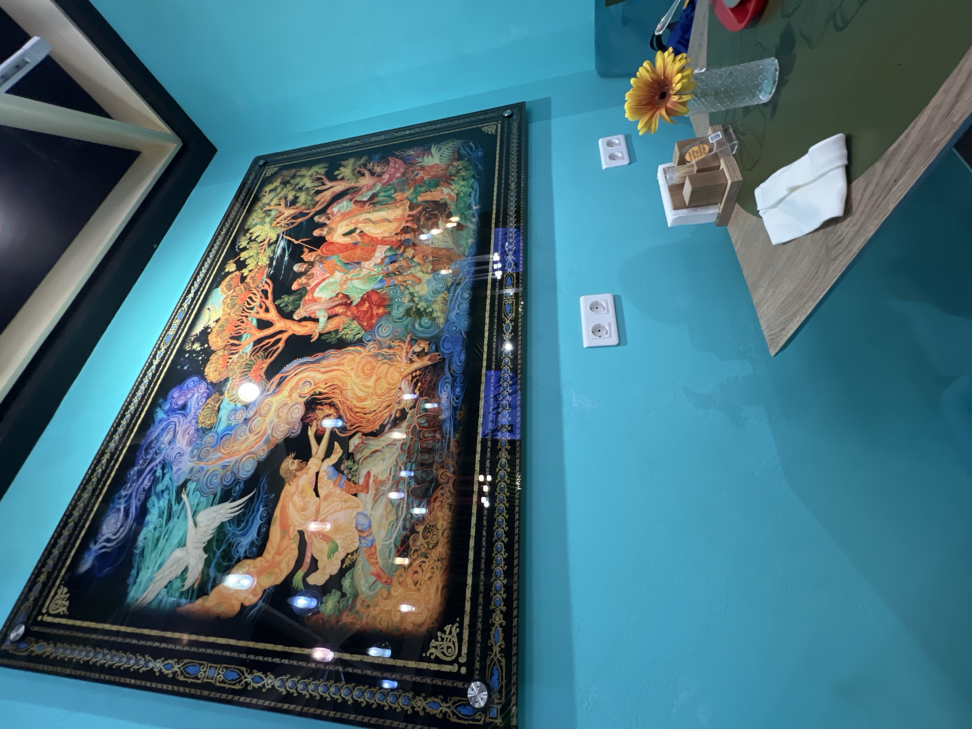 A framed, ornate painting hangs on a bright turquoise wall, depicting a fantastical scene with mythical figures and creatures rendered in vibrant colors. A bird with outstretched wings soars through swirling blue and green clouds above a figure draped in flowing orange fabric. Below, a group of people in colorful attire gather amongst trees. The painting is illuminated by small, embedded lights that reflect off its surface.
On the table below the painting, a green tablecloth covers the surface. A clear glass vase holds a single yellow gerbera daisy. Nearby, a napkin holder with toothpicks and small paper packets is positioned next to a white folded napkin. Two electrical outlets are visible on the turquoise wall.
The scene suggests an interior space, likely a cafe or restaurant, possibly in Moscow, Russia, given the artistic style of the painting which resembles traditional Russian folk art. The lighting is dim, with the overhead ceiling visible in the upper right corner, featuring dark panels and light-colored beams. The overall impression is one of a decorated interior space with a focus on a prominent artwork.