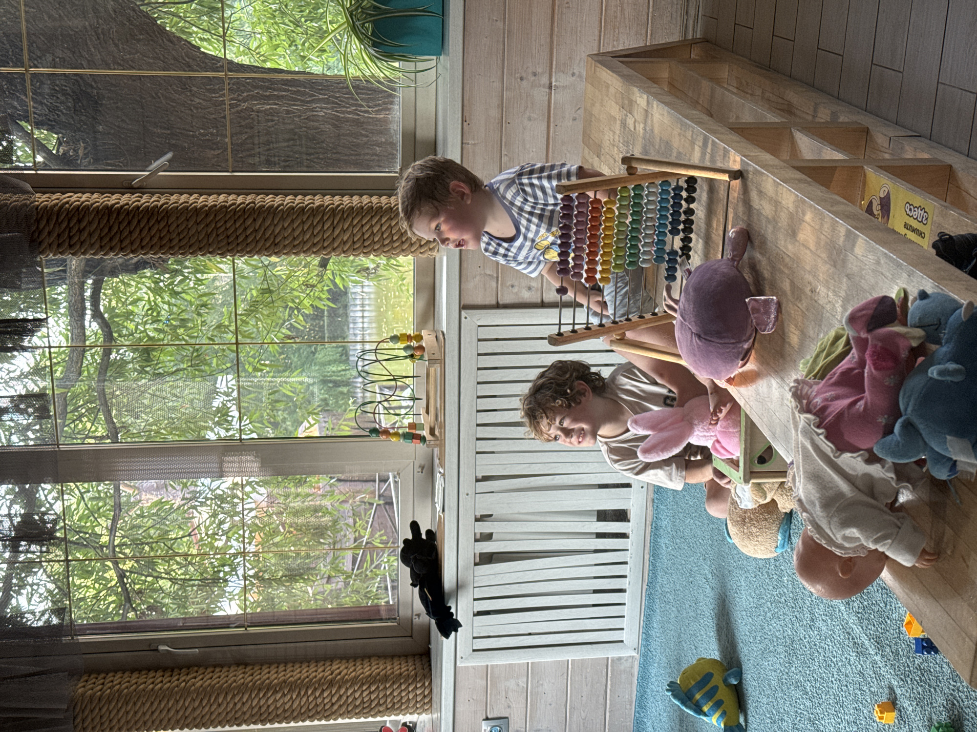 The image depicts an indoor play area, likely within a residence in Sovkhoz imeni Lenina, Russia, due to the presence of a poster with Russian text visible on a shelf. Two young children are present, one boy with blonde curly hair sitting on a blue rug and looking upwards, and another boy wearing a striped t-shirt also looking upwards. The environment is a room with wooden paneling on the walls. Large windows offer a view of lush green trees and water, suggesting an outdoor natural setting beyond the building.
In the foreground, a wooden abacus with colorful beads is placed on a wooden table. Various toys are scattered around the children and on the rug, including a yellow and blue fish toy, a pink bunny stuffed animal, a brown teddy bear, a doll with pink pants and a white shirt, and other plush toys and building blocks. A black stuffed animal is visible on a white radiator positioned beneath the window. The overall scene suggests a domestic and playful environment.
