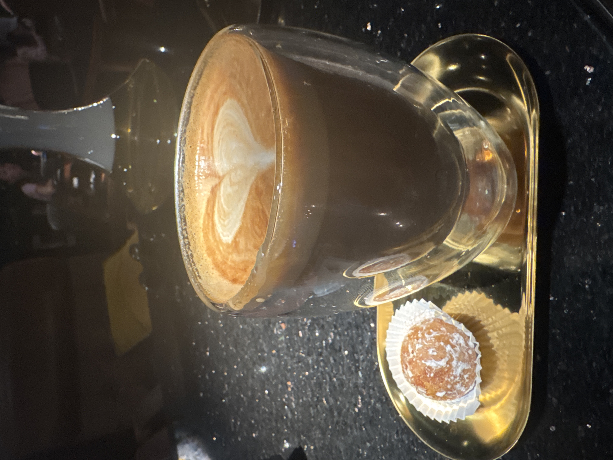 The image, rotated 90 degrees counter-clockwise, captures a close-up, high-angle view of a coffee drink and a small pastry presented on an elegant tray. The scene is set indoors, likely within a modern cafe or restaurant, possibly in North Kuta, Indonesia, given the general ambiance suggested by the blurred reflections in the dark background.
The primary subject is a clear, double-walled glass mug holding a latte or cappuccino. The surface of the coffee is adorned with intricate white latte art, forming a distinct heart shape, which stands out against the rich, frothy brown layer. The dark brown coffee liquid is visible through the transparent glass.
To the right of the coffee, on a small, lustrous golden metallic tray, sits a single, round pastry. It is cradled within a white, fluted paper liner and generously dusted with white powdered sugar, suggesting a sweet treat like a mini donut or a truffle. The tray has a sleek, slightly curved design, complementing the presentation.
Both the coffee and pastry rest on a dark, speckled surface, resembling polished black granite or quartz, which reflects subtle points of light from its sparkling inclusions. The background is softly out of focus, revealing indistinct shapes and reflections, possibly indicating other guests or interior furnishings. No text is visible in the image. The overall atmosphere conveyed is one of a comfortable and stylish setting for a coffee and dessert experience.
