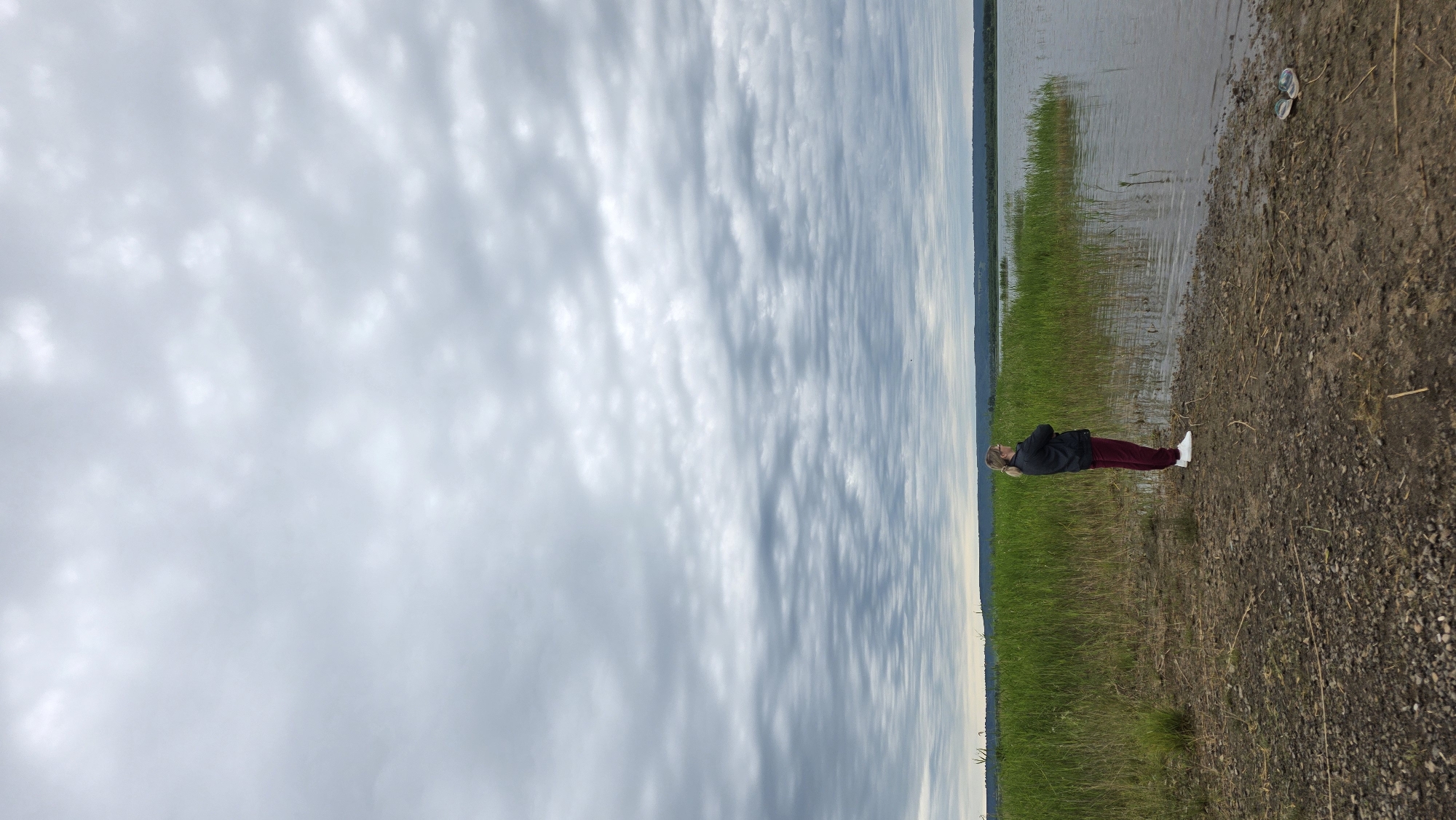 A person stands on the shore of a body of water, facing away from the camera. The person is wearing a dark jacket and dark red pants, with white shoes. They have their arms crossed. Behind the person is a thick growth of tall, green grass. The body of water appears to be a lake or slow-moving river, with the far shore visible in the distance. The sky is overcast with a pattern of layered, grey clouds. The ground in the foreground is composed of dirt and small pebbles. The scene is likely located in the village of Kupoyarovo, Russia, given the natural landscape.