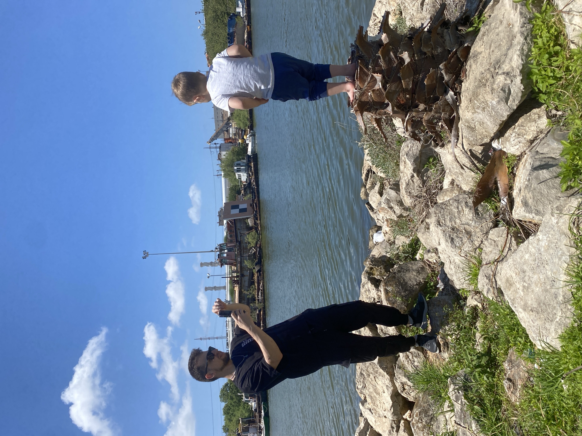 A man in sunglasses, a black t-shirt, and black pants is standing on a rocky bank, holding a phone and seemingly taking a picture of a young boy. The boy, wearing a white t-shirt and blue shorts, is standing with his feet on the base of a palm tree trunk at the edge of the rocky bank. The scene is by a body of water, potentially a river or harbor, with industrial buildings and cranes visible in the background. The sky is blue with scattered clouds. This is likely a location in Haifa, Israel, near the port or industrial area along the waterfront.