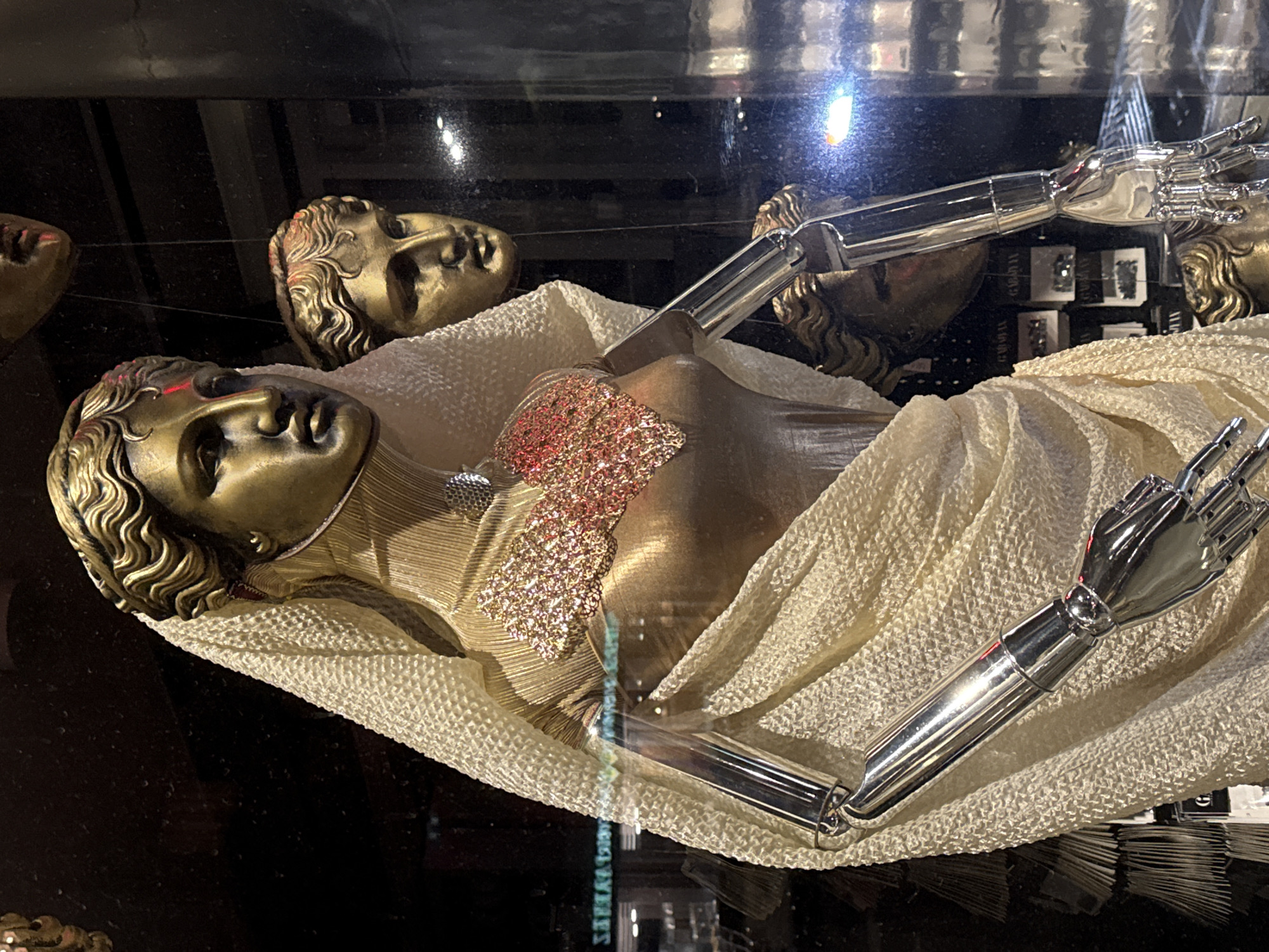 A display case in Yerevan, Armenia, contains a bronze statue of a reclining figure with a human-like face and wavy hair. The figure is draped in a textured, white cloth. Two silver, robotic arms extend from the statue's body. Scattered bronze face-like sculptures are arranged in the background.
