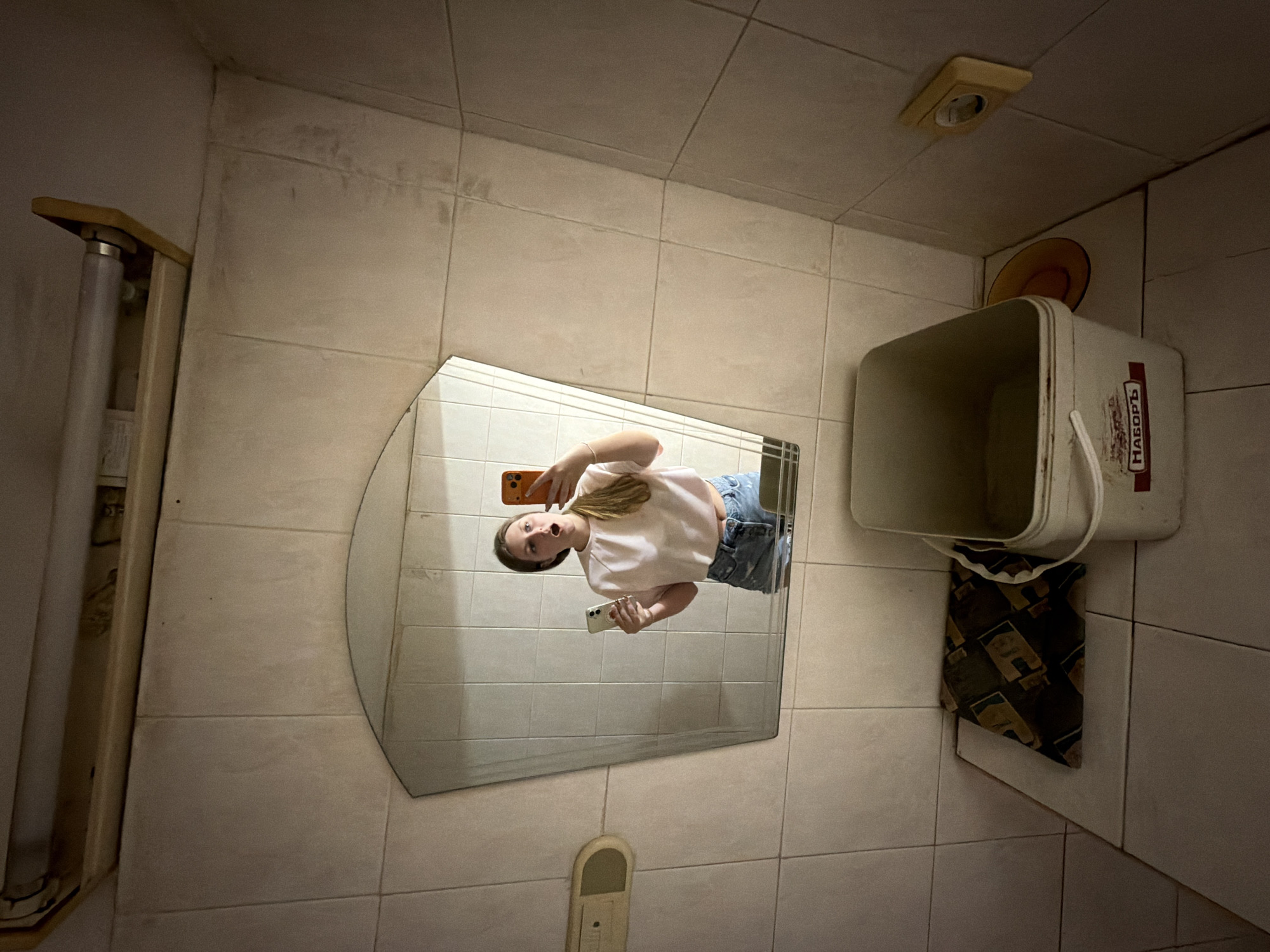 A young woman stands in a tiled bathroom, holding two smartphones and taking a selfie in a mirror. She is wearing a light pink crop top and denim shorts. To her right, a white bucket with the Cyrillic word "НАБОРЪ" (nabory, meaning "set" or "kit") is visible on a tiled surface, next to a patterned cloth. Above the mirror, a fluorescent light fixture is attached to the wall. The bathroom appears to be in a public facility, possibly a rest stop or public restroom, given the utilitarian tiling and the presence of the bucket. Without specific visual cues linking the scene to Burgas, Bulgaria, its location cannot be confirmed. The environment is dimly lit, with harsh fluorescent lighting.