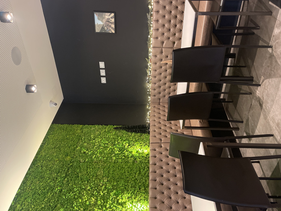 This image depicts the interior of what appears to be a modern restaurant, cafe, or lounge in Sondrio, Italy. The room is empty of people, with a focus on its contemporary decor and seating arrangements.
On the left side of the frame, a vibrant, bright green wall covered in artificial moss or a similar plant material dominates the lower half, illuminated from below to create a glowing effect. Above this, the wall transitions to a dark grey or black, and further up, a white perforated ceiling with integrated spotlights is visible. A small security camera is mounted where the white ceiling meets the dark wall.
The back wall is dark grey or black, featuring a single framed picture hanging centrally; it appears to be the iconic black and white "Abbey Road" album cover by The Beatles. Below the picture, three white electrical outlets or switches are vertically stacked on the wall. Along the top edge of a long, tufted banquette that runs parallel to the back wall, delicate string lights or fairy lights are draped, adding a warm, decorative touch.
To the right, several tables with white tops and dark bases are arranged in front of the banquette. Black chairs with sleek designs and dark frames are pulled up to these tables. The floor is a light grey, possibly concrete or tiled, providing a neutral base for the furniture.
The overall atmosphere is clean and modern, characterized by contrasting textures and colors, including the plush brown banquette, the organic green moss wall, and the stark black and white elements. The interior lighting, including spotlights and decorative string lights, suggests an indoor setting, potentially during the evening. No specific activity is taking place, and there is no visible text to read within the image.