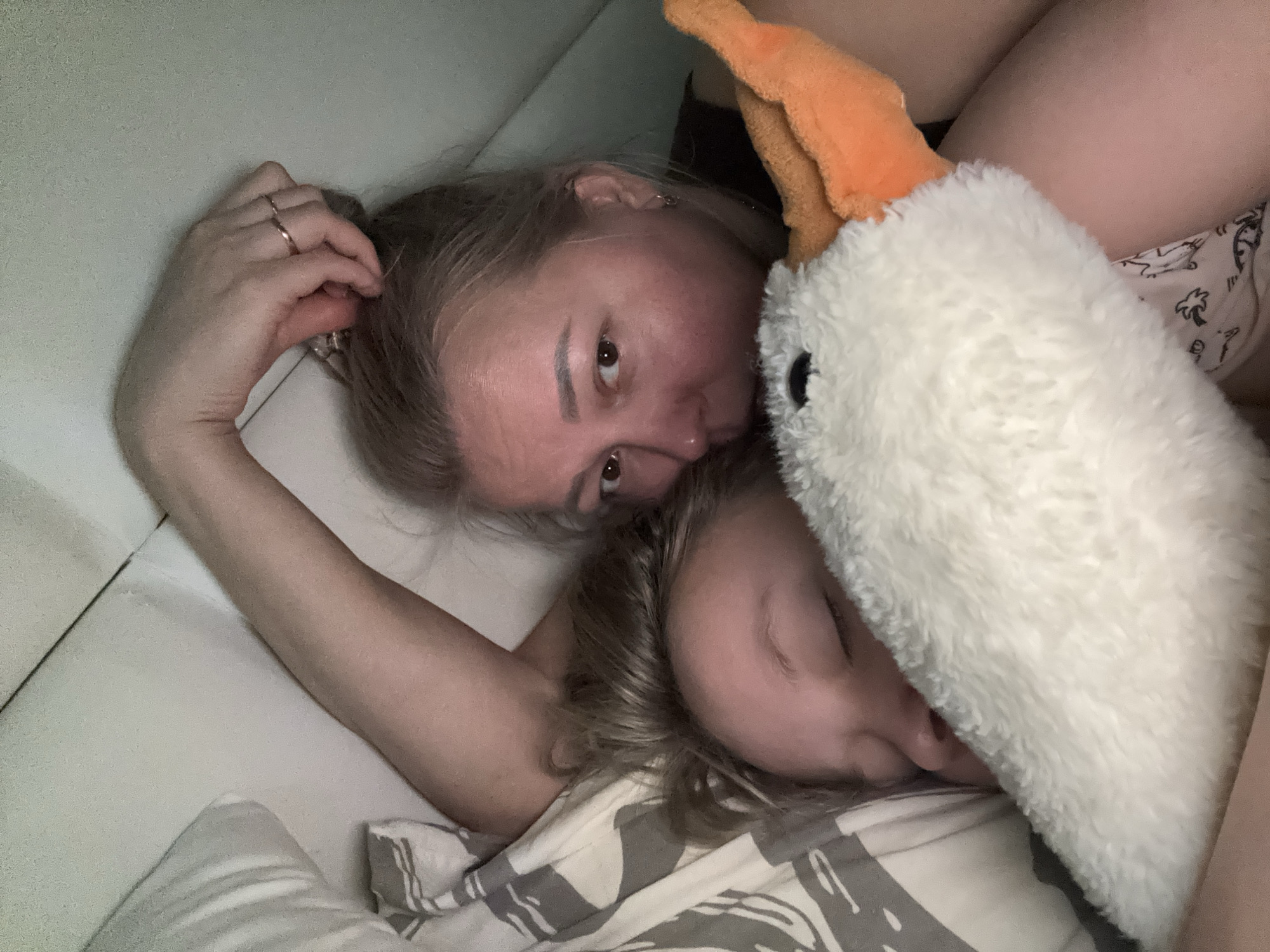 A woman and a child are lying in bed, with the child's head near the woman's chest and shoulder. The woman is looking at the camera, while the child's eyes are closed. A white plush toy with an orange beak is next to the child. The background appears to be a bedroom. The location is unknown, but a reference to the city of Izhevsk, Russia, is required.