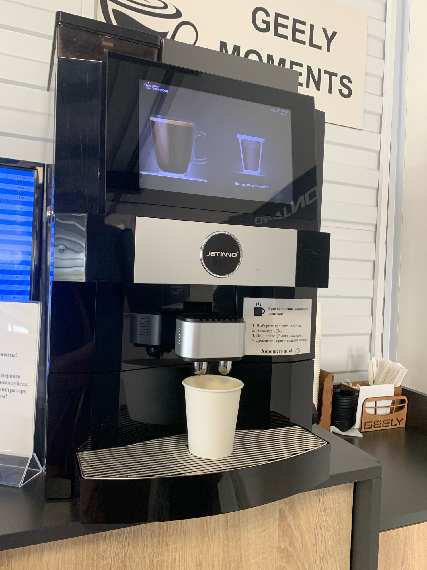 In the city of Chyzhewicki, Belarus, a modern black coffee machine is positioned on a counter. The machine features a digital display showing a cup of coffee and the text "Ваш напиток готовится" (Your drink is being prepared). Below the screen, the brand name "JETINNO" is visible. Instructions for preparing coffee are displayed on a sticker, which include steps to select a drink, press "OK," scan a QR code, and wait for preparation. A white paper cup filled with coffee sits in the dispenser, ready for use. To the right of the coffee machine, a wooden holder with napkins and a black container are visible, with the "GEELY" logo on the holder. The overall environment appears to be a clean, modern establishment, likely a cafe or a business offering complimentary beverages.