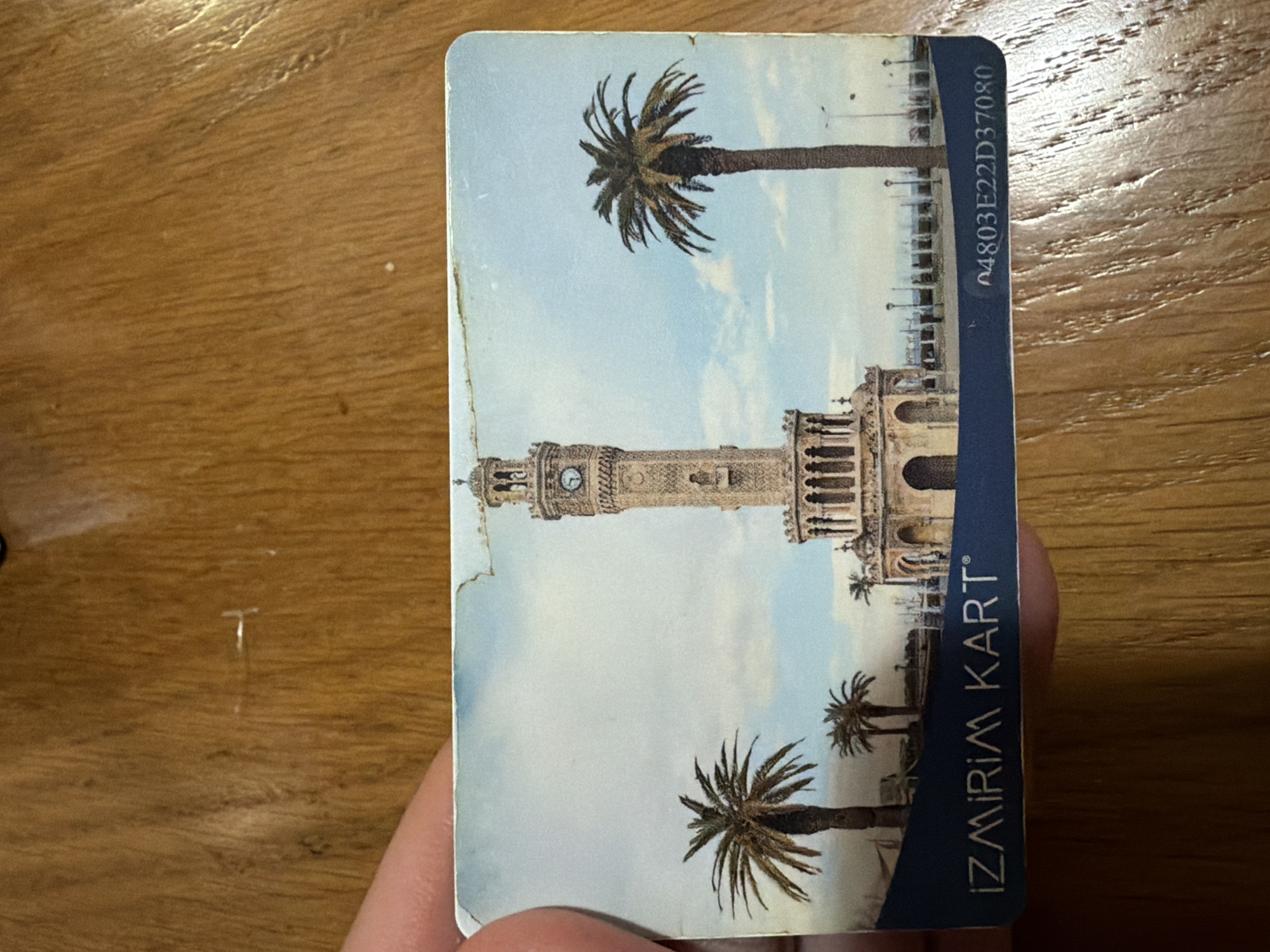 The image shows a worn "Izmirim Kart" card featuring the Izmir Clock Tower in Izmir, Turkey, and palm trees. The card has the serial number "n4803E22D37080" printed on its edge. The card is resting on a wooden surface, and a person's hand is holding the bottom edge.