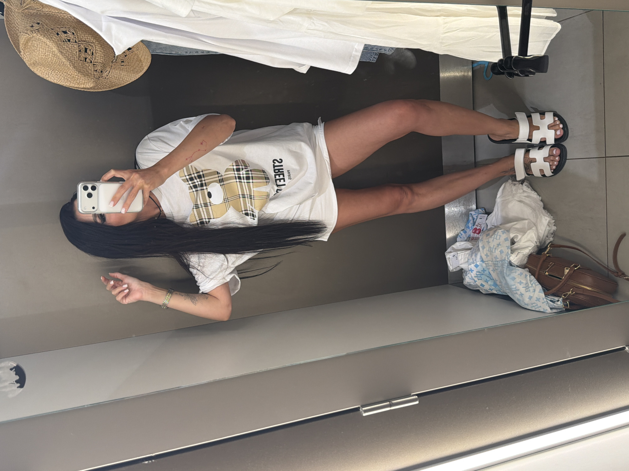The media file depicts a person taking a mirror selfie in what appears to be a fitting room or a changing area. The person is standing and holding a white smartphone in their left hand, angled to capture their reflection. They are wearing a loose white t-shirt with a graphic of a bear in a plaid pattern and a pair of white slide sandals. Their long, dark hair is visible, framing their face. Visible on their arm is a tattoo. The background shows a wall with a reflective surface (the mirror), and on a shelf to the right, there are some folded clothes and a brown handbag. Above the person, a straw hat is hanging, and white fabric is draped. The setting appears to be indoors, possibly in a retail environment given the fitting room aesthetic. The lighting suggests daytime. The overall tone is casual and self-documenting. The image does not contain any visible text.