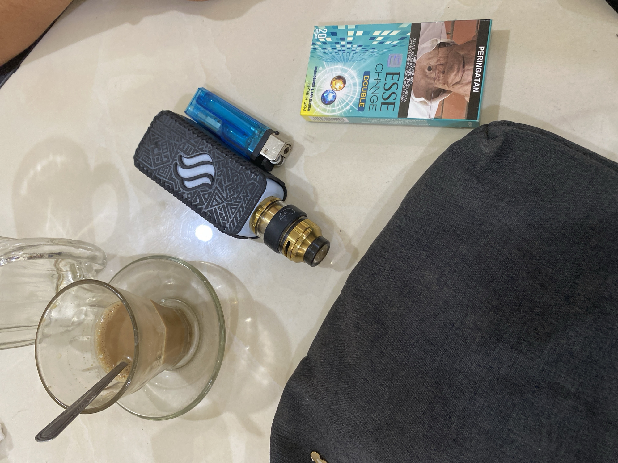 A flat lay image captures a tabletop scene in Lhokseumawe, Indonesia, populated with a mix of items associated with daily habits and consumption. A glass cup filled with a light-brown liquid sits on a clear saucer, a spoon resting inside. Adjacent is a black and gold vaping device, accompanied by a blue lighter. Next to these items, a pack of Esse Change Double cigarettes is present. Part of a dark fabric, likely clothing or a bag, occupies a significant portion of the right side of the frame. The environment suggests a casual setting, possibly a café or home table.