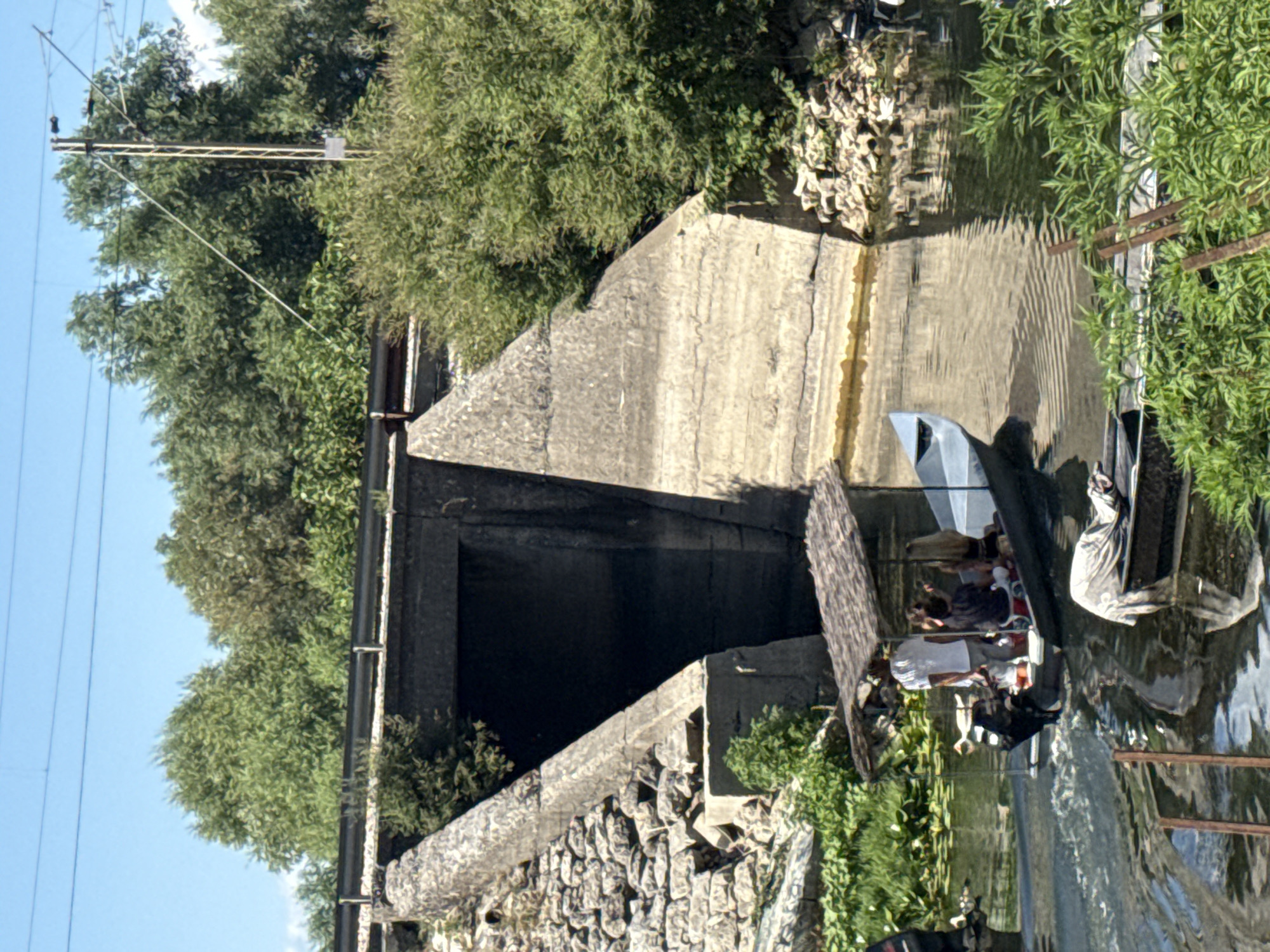 The image depicts a canal with a boat carrying several people, passing under a concrete bridge that appears to be part of an elevated railway or road infrastructure. On the left side of the canal, there is abundant green vegetation. The concrete structure shows signs of age and weathering, with stones and debris at its base. The water is calm, with reflections of the surrounding trees and the bridge. A second, smaller boat is visible in the foreground, partially covered with a grey cloth. In the background, lush green trees dominate the landscape, and overhead power lines are visible against a clear blue sky, suggesting an outdoor, possibly rural or semi-rural, setting within Vranjina, Montenegro.