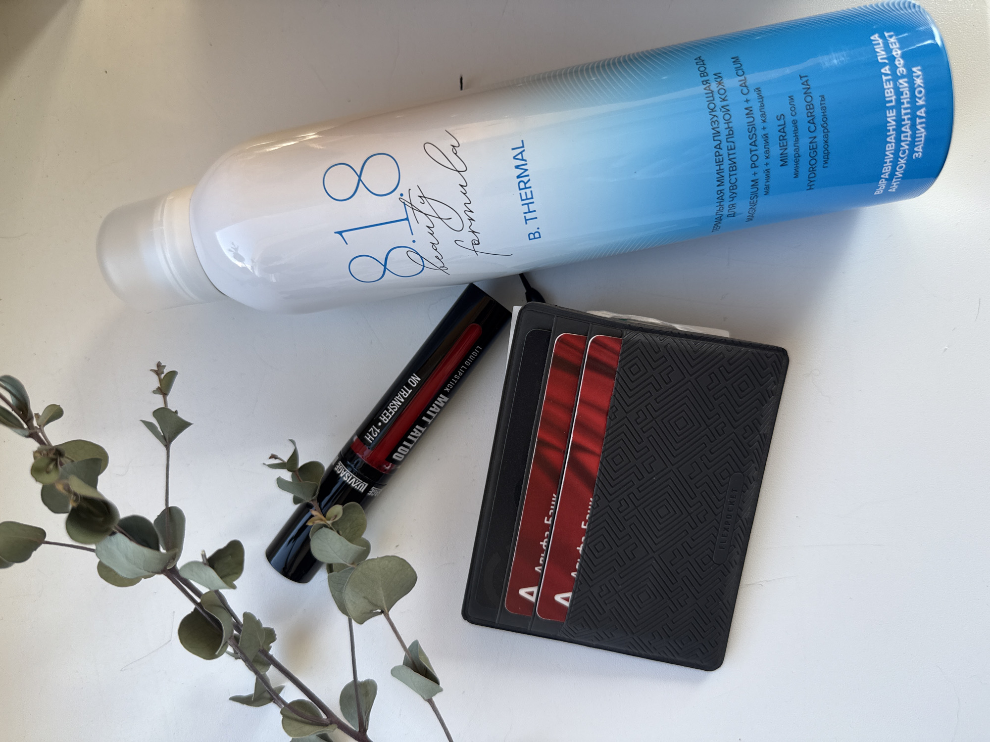 This overhead flat lay photograph, taken in Izhevsk, Russia, showcases an arrangement of personal care items and a wallet on a light, neutral surface. The bright lighting suggests it was taken during the day.
The most prominent item is a large, white and blue spray bottle, likely a facial mist or thermal water. Its label clearly displays "818 beauty formula B. THERMAL." Significant Russian text is also visible, identifying it as "ТЕРМАЛЬНАЯ МИНЕРАЛИЗУЮЩАЯ ВОДА ДЛЯ ЧУВСТВИТЕЛЬНОЙ КОЖИ" (THERMAL MINERALIZING WATER FOR SENSITIVE SKIN). It lists ingredients such as "MAGNESIUM + POTASSIUM + CALCIUM," "MINERALS," and "HYDROGEN CARBONAT." The benefits highlighted include "ВЫРАВНИВАНИЕ ЦВЕТА ЛИЦА" (FACE COLOR EVENING), "АНТИОКСИДАНТНЫЙ ЭФФЕКТ" (ANTIOXIDANT EFFECT), and "ЗАЩИТА КОЖИ" (SKIN PROTECTION).
Next to the bottle is a black tube of "LUXVISAGE LIQUID LIPSTICK MATT TATTOO," specifying "NO TRANSFER 12H." The visible part of the tube is dark, with red accents.
Below these items lies a sleek, black textured wallet, which features the text "FLEXPOCKET" on its side. Three red cards are partially visible, tucked into the wallet, adding a pop of color. A sprig of dried eucalyptus with muted green leaves is positioned in the lower-left corner, adding a natural, decorative element to the composition. No activity is taking place; the items are static, arranged for display.