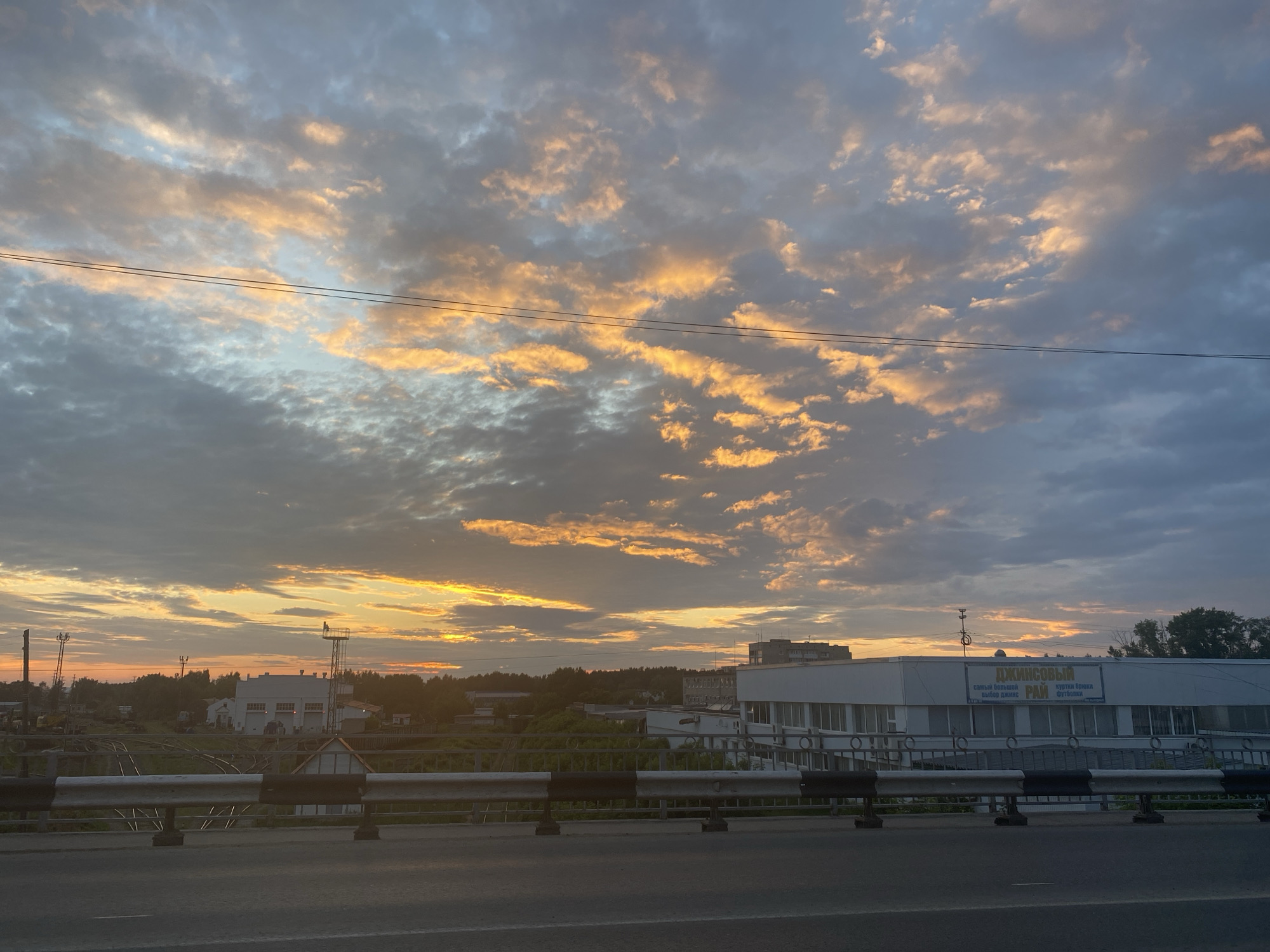 An outdoor medium shot captures a sunrise over an industrial area, with the sky filled with dramatic clouds reflecting the warm hues of the setting or rising sun. The clouds are a mix of gray, blue, and vibrant orange and yellow streaks. Two thin, dark wires stretch horizontally across the upper portion of the frame. In the middle ground, a low-lying white building with numerous windows is visible, bearing a sign in Russian that appears to advertise denim clothing and outerwear. This building is part of a complex that includes other industrial structures and what looks like railway tracks. The foreground features a concrete road or bridge deck, with a safety barrier composed of alternating black and white reflective panels. The surrounding landscape is dotted with trees and other buildings, suggesting a transition zone between urban development and nature. The overall scene is characterized by the interplay of natural light and the built environment.