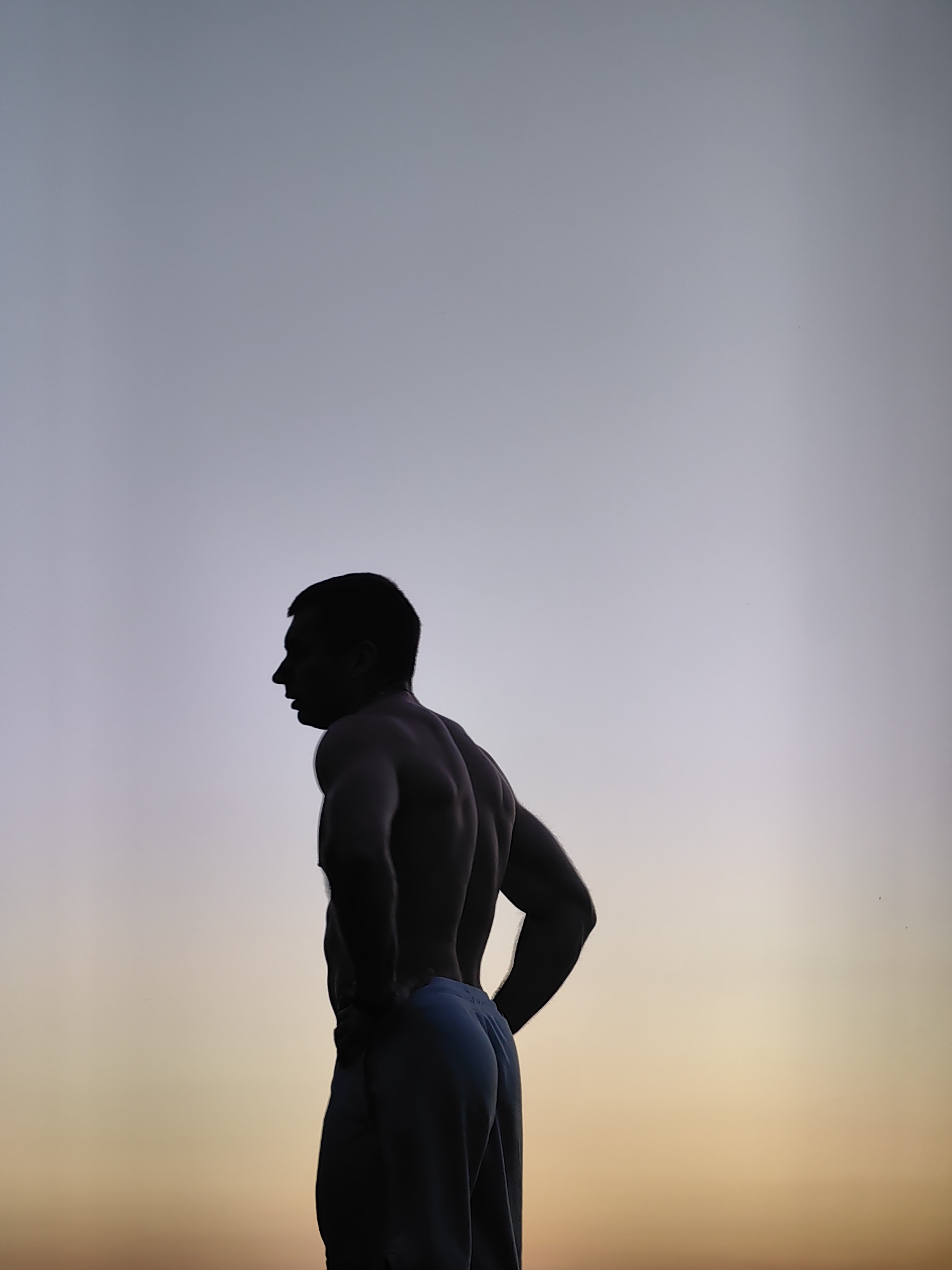 A silhouette of a shirtless muscular man is visible against a gradient sky that transitions from pale blue at the top to warm orange and yellow hues towards the bottom, suggesting either sunrise or sunset. The man is positioned facing left in profile, with his hands on his hips and his head slightly turned away from the viewer. His musculature, particularly his back and shoulders, is well-defined in the silhouette. He appears to be wearing light-colored pants. The image is cropped to focus on the man and the sky, with no identifiable urban or natural landscape elements visible. The specific location in Varna, Bulgaria, is not discernible from the visual information provided in the image.