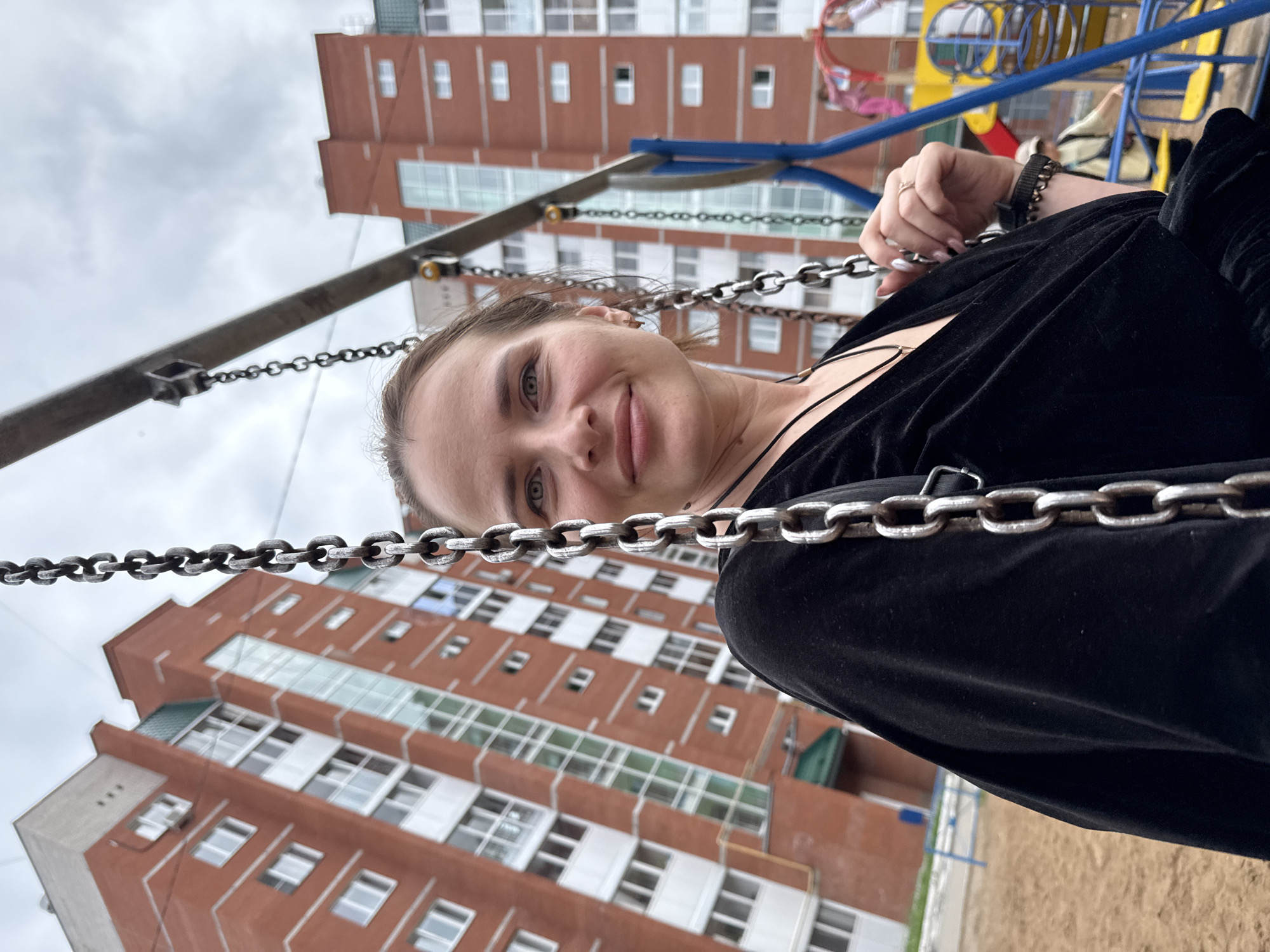 A woman with light brown hair, wearing a black top, is seated on a swing. She is looking towards the camera with a slight smile. In the background, a modern brick apartment building dominates the scene. The building has multiple floors with numerous windows and a distinctive green sloped roof. The setting appears to be a playground, with sand on the ground and parts of playground equipment visible, including blue poles and yellow seating. The sky is overcast, suggesting a cloudy day. The location is identified as the city of Chaikovsky, Russia.