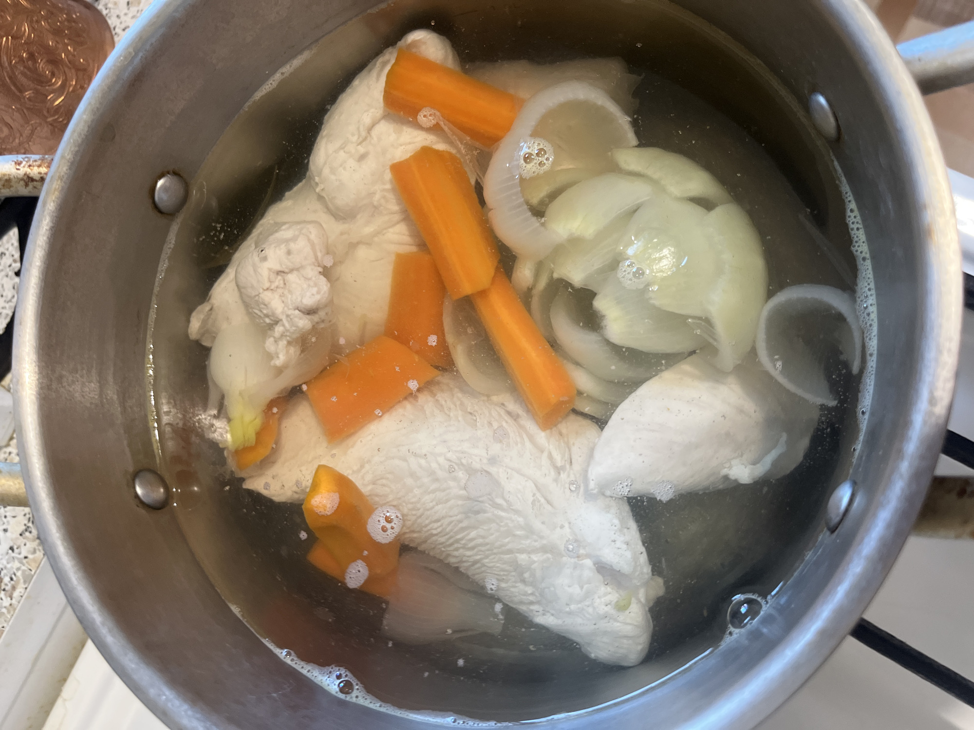 A top-down, close-up shot shows chicken pieces, carrots, and onions simmering in a pot of clear broth. The chicken is white and appears cooked, with some pieces showing a stringy texture. The carrots are cut into thick rounds, and the onions are sliced into large segments. Bubbles are visible on the surface of the broth, indicating it is at a simmer. The pot is a silver-colored metal with visible rivets on the rim. The background is blurred and suggests a kitchen environment with speckled countertops. The lighting is bright, highlighting the contents of the pot.