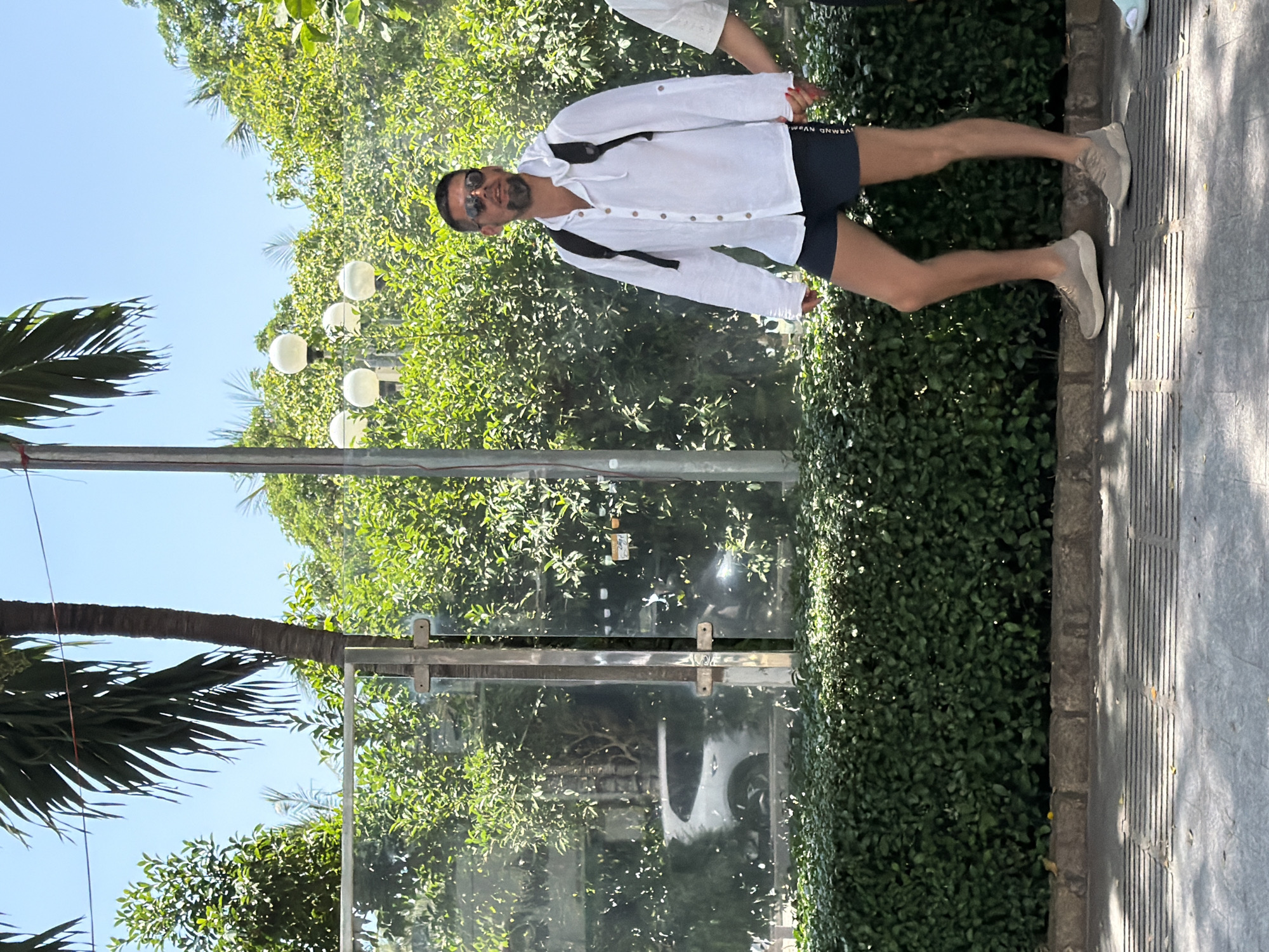 The image depicts a man walking outdoors in a city setting, likely Nha Trang, Vietnam, given the context. He is wearing a white, hooded, long-sleeved linen shirt, dark blue shorts, sunglasses, and light-colored athletic shoes. A black backpack is visible on his shoulders. He is walking on a paved sidewalk adjacent to a manicured green hedge. Behind the hedge, a glass partition is visible, possibly part of a building or bus stop, reflecting trees and a white vehicle. Further in the background, lush green foliage and palm trees are present under a clear blue sky. The man appears to be walking casually, and another person's hand holding his is slightly visible on the right side of the frame. The scene suggests a bright, sunny day.