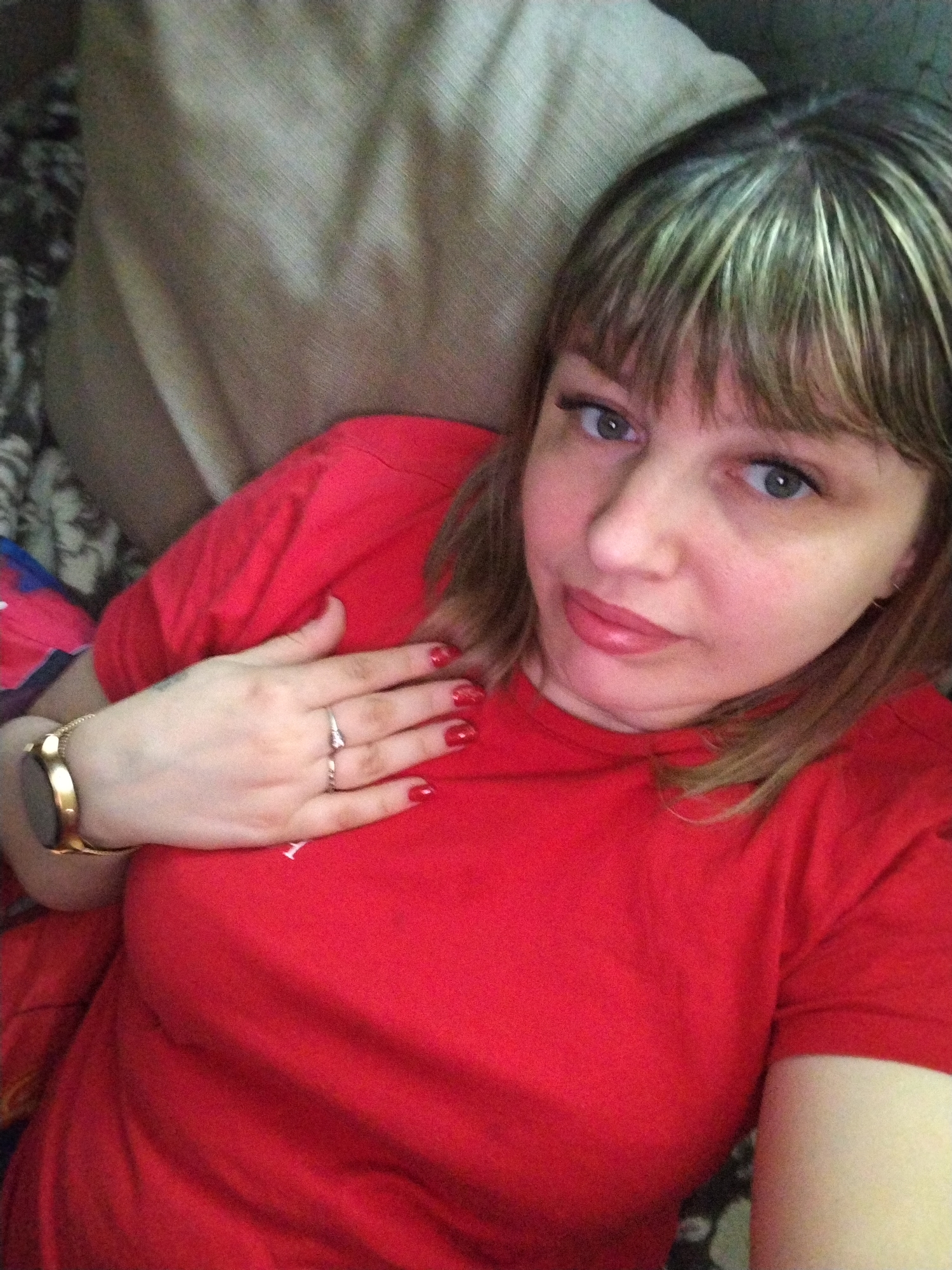A woman with blond-highlighted bangs is lying down. She wears a red t-shirt, gold watch, and two rings on her left hand. Her nails are painted red. Behind her is a beige pillow and a patterned surface. No location is evident in the image.