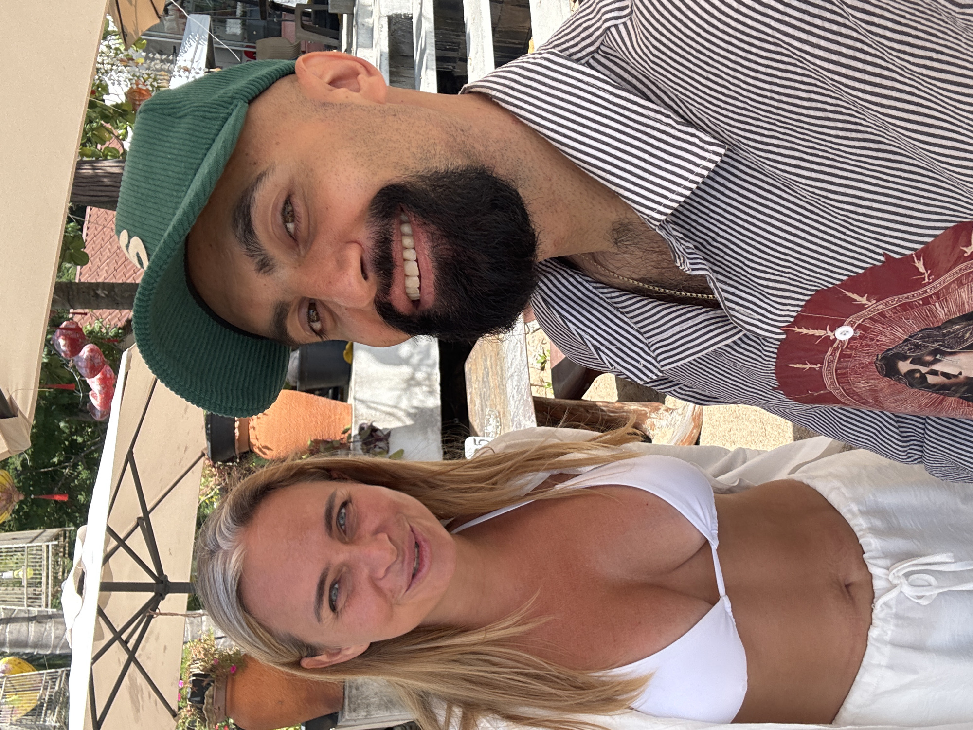 A medium shot shows a man and a woman posing for a selfie outdoors on a sunny day. The man, positioned on the right, is wearing a green corduroy baseball cap, a black and white striped button-down shirt, and a gold chain necklace. He has a beard and is smiling at the camera. The woman, on the left, has blonde hair and is wearing a white bikini top and a white semi-sheer cover-up. She is also smiling. In the background, there are outdoor umbrellas, potted plants, and what appears to be stairs leading up to a building, suggesting a tropical resort or restaurant setting. The overall scene conveys a casual and relaxed atmosphere. The location is the city of Ko Samui District, Thailand.