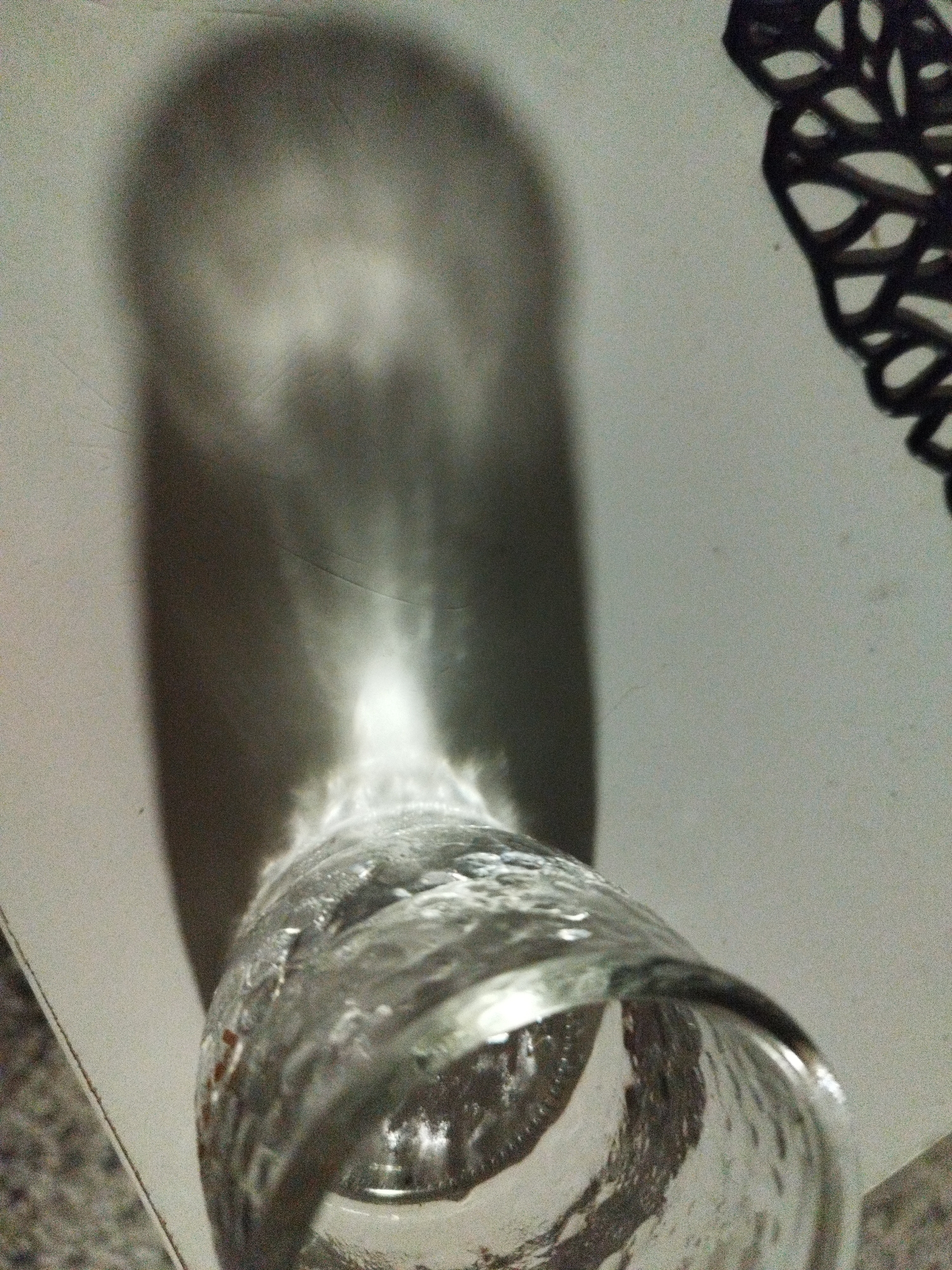 The image depicts a clear glass jar casting a shadow on a light-colored surface. The jar is positioned near the bottom of the frame, angled upwards. Light is passing through the jar, creating a concentrated bright spot in the shadow. A dark, intricate object is visible in the upper right corner of the image. The photograph appears to be taken indoors. There is no information to confirm the location is Sofia, Bulgaria, nor any people, subjects, objects, actions, or interactions beyond the description provided by the image itself.