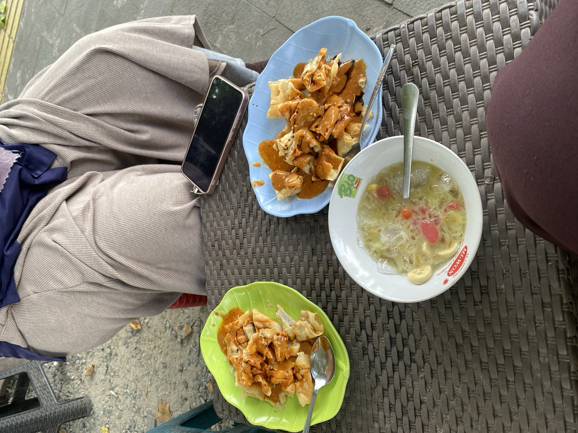 An overhead shot shows a table with three dishes of food and a cell phone. The table is covered with a woven brown placemat. In the foreground, a person's legs are visible, wearing dark maroon pants. To the left is a bright green leaf-shaped plate filled with fried food covered in a reddish-orange sauce, with a silver spoon resting on the plate. To the right of the green plate is a light blue leaf-shaped plate with a similar fried food dish, also covered in a reddish-orange sauce. In the center of the table, closer to the viewer, is a white bowl filled with a clear liquid, ice, sliced bananas, and red jelly-like pieces. A silver spoon is placed in this bowl. A black smartphone with a rose gold case lies on the table, just above the blue plate. In the background, a person is seated, wearing a beige ribbed garment and a dark blue headscarf. The setting appears to be an outdoor eating area, with a sandy ground and some fallen leaves visible behind the table, possibly in a roadside stall or warung in Banjarbaru, Indonesia.