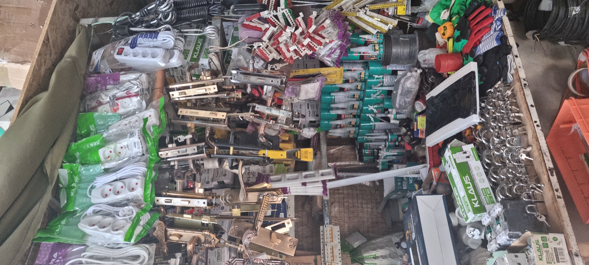 This image depicts a retail stall in Bokhtar, Tajikistan, likely a hardware or general store. The stall is densely packed with merchandise, primarily hardware items and electrical accessories. Prominently displayed are numerous door handles, locks of various sizes with keys, and electrical extension cords with multiple sockets.
On shelves and hanging, a variety of other items are visible, including what appear to be tubes of adhesive or sealant, electrical circuit breakers in packaging, and bundles of wire or cable. The overall environment suggests a busy, functional retail space, with items arranged in a manner to maximize visibility and accessibility for customers. The lighting appears to be natural or from overhead fixtures, casting some shadows but generally illuminating the products.
