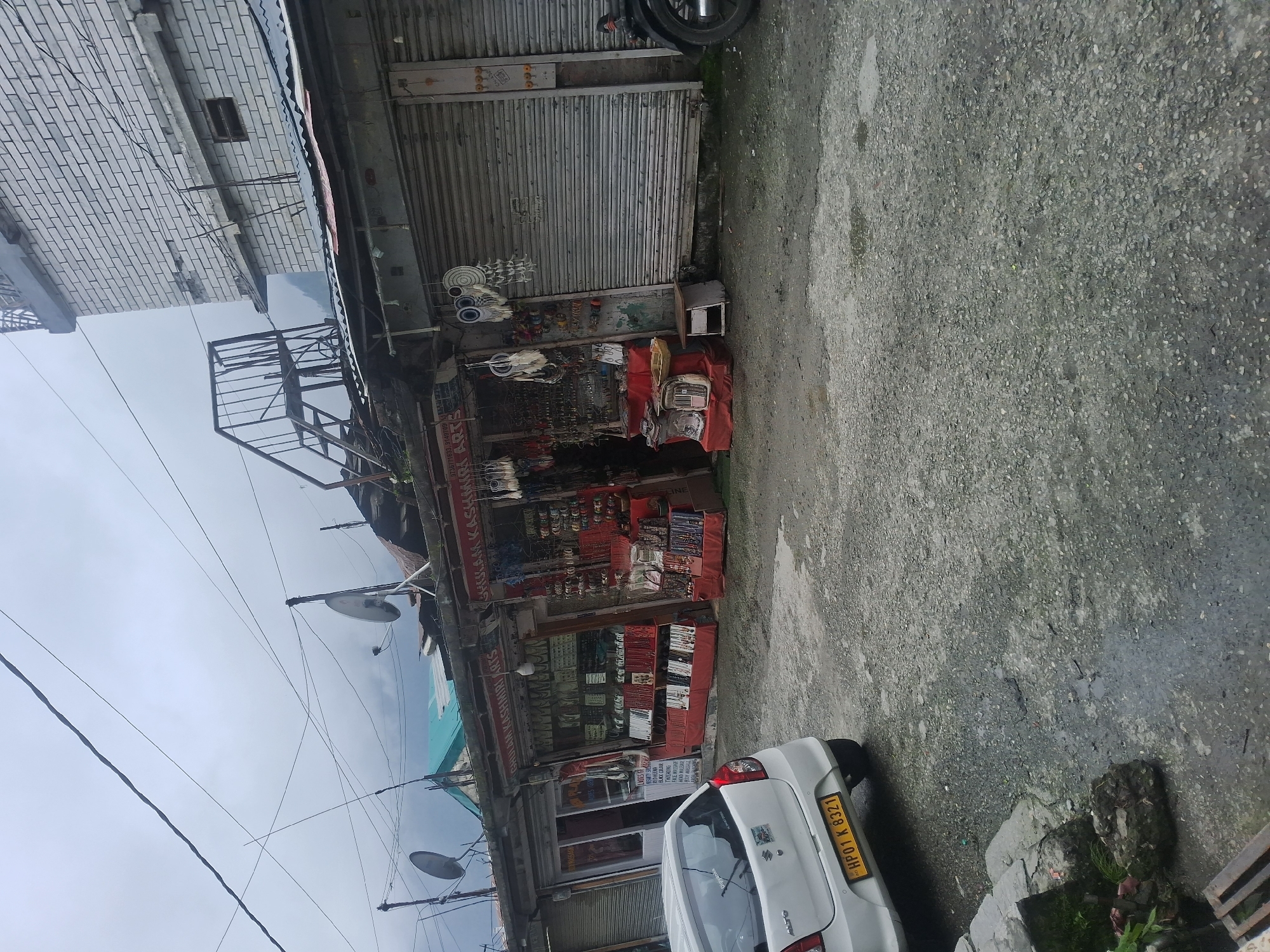 The image depicts a street scene in Vashisht, India, characterized by a row of small shops. A white car with a yellow license plate, "HP01 K 8321", is parked on the left side of the frame, partially obscuring the pavement. The shops are adorned with various items, including dreamcatchers, jewelry, and other handicrafts, suggesting they are souvenir or artisan shops. Some of the shop shutters are down, indicating they might be closed. An outdoor staircase leading to an upper level is visible on the left side of the buildings. The environment appears to be a cloudy day, with the shops situated along a somewhat uneven, gravelly road. The architecture suggests a local, possibly traditional, style with corrugated metal roofing and brickwork on the upper floors of some buildings. The overall impression is of a bustling market area, potentially in a tourist-frequented location.